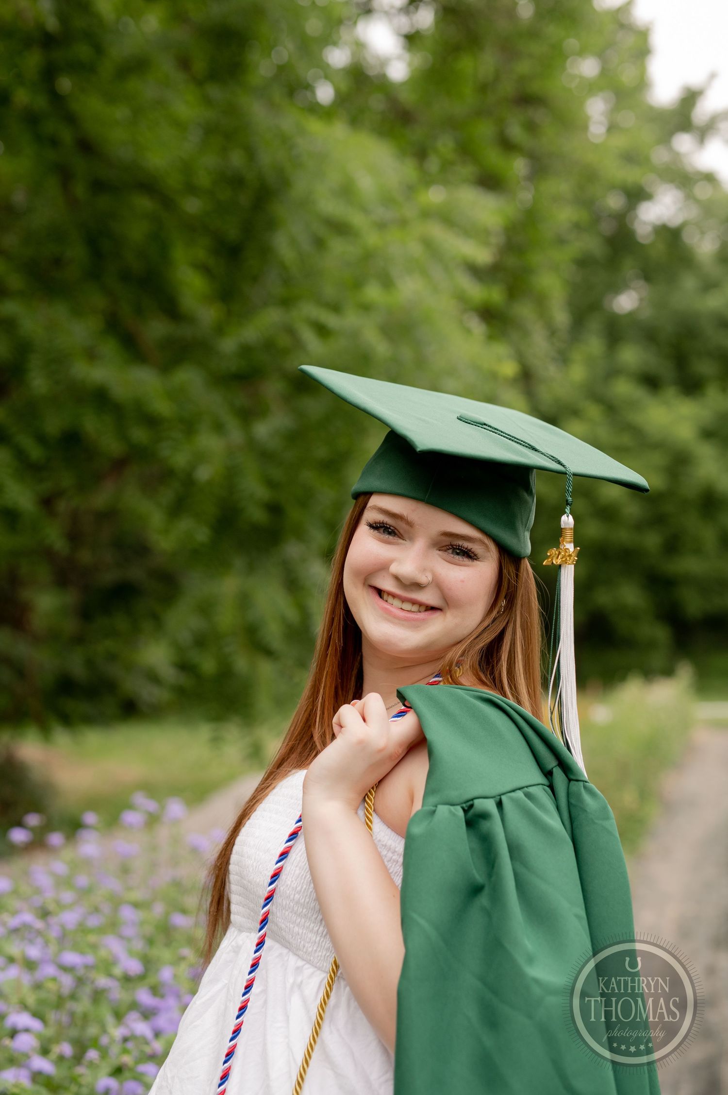 Graduate wearing green cap with tassel and white dress smiles against garden backdrop.