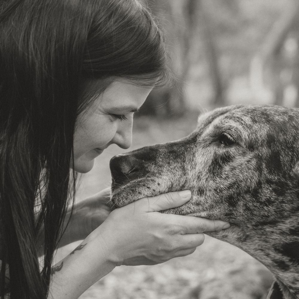 Black and white photo of a woman tenderly holding a dog's face close to hers in an affectionate moment outdoors.