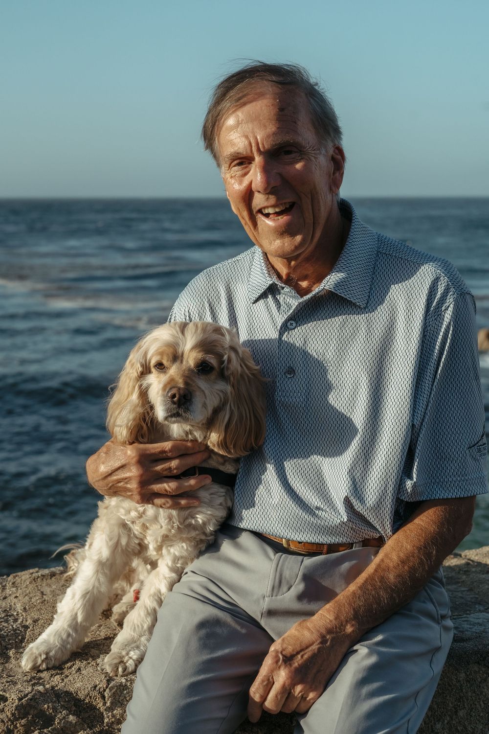 Senior man in grey polo shirt sits with his Cocker Spaniel dog on rocky beach with ocean waves in background.