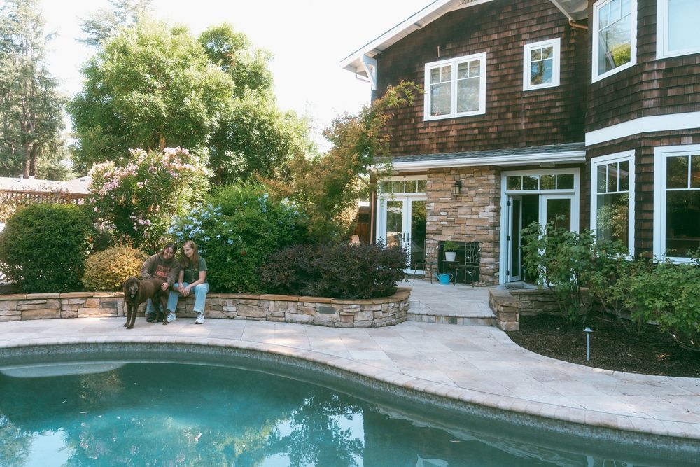 Luxurious backyard pool area with a brown shingle home, stone patio, lush greenery, and people relaxing near the water.