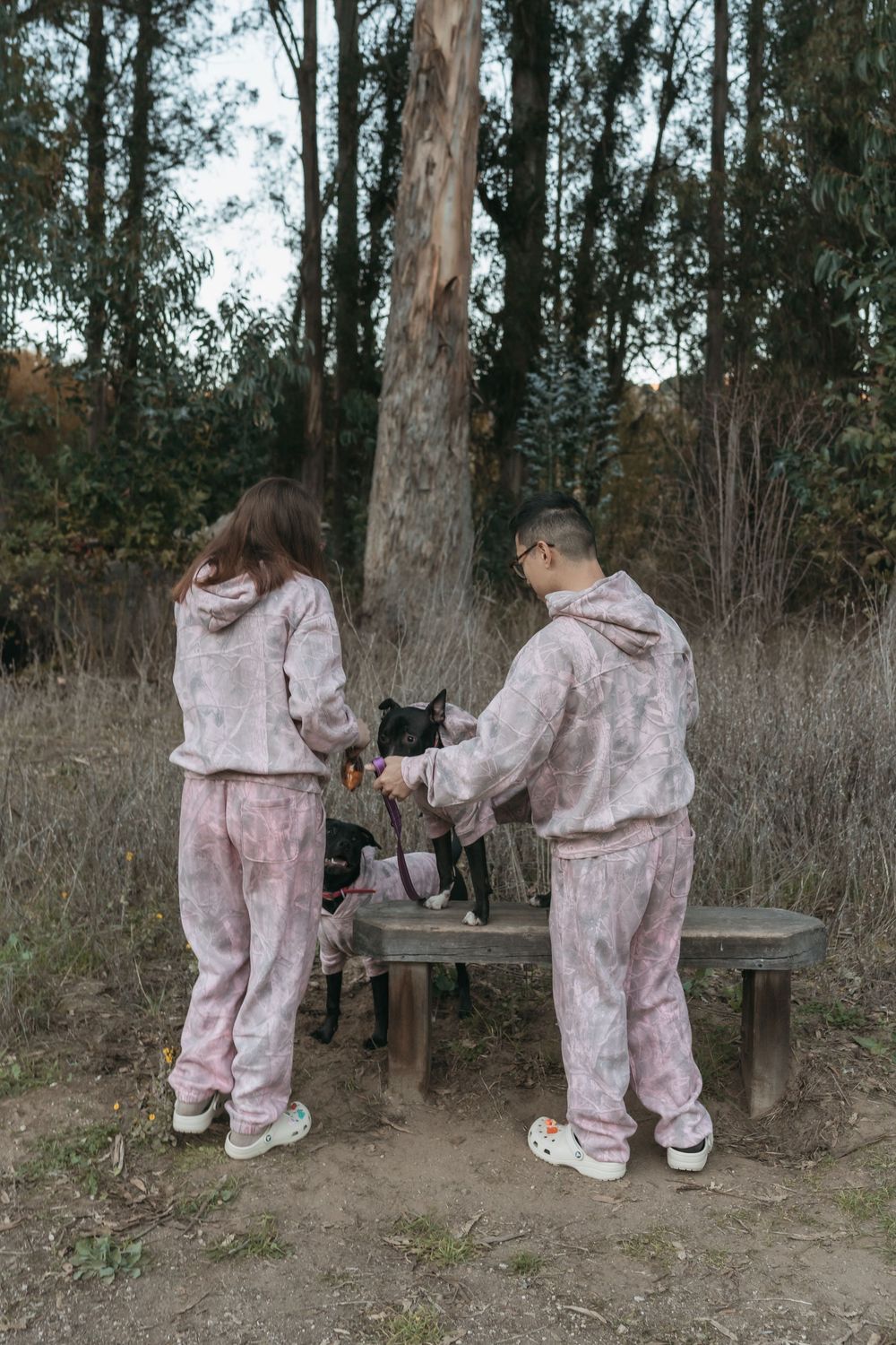 Two people in pink plush onesies interact with a black cat on a wooden bench in a wooded outdoor setting.