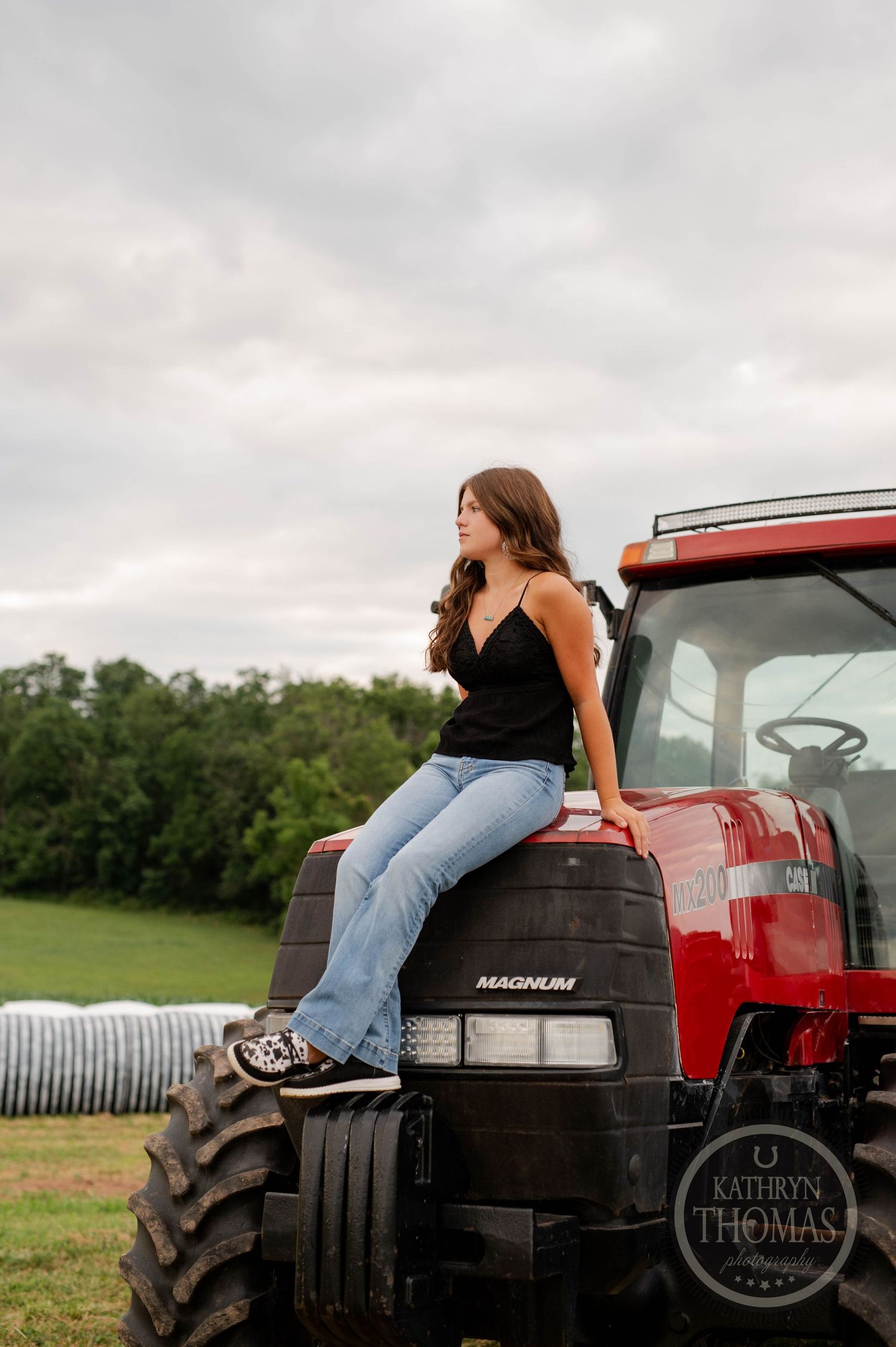 Woman in casual attire sits on red tractor hood against cloudy rural backdrop.