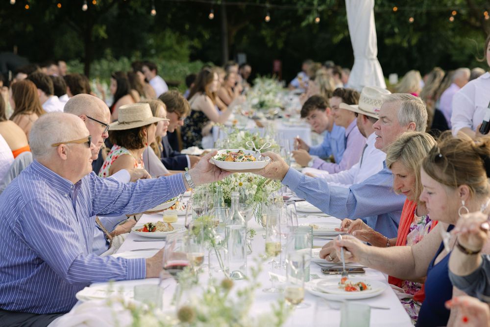 Guests seated at a long outdoor table enjoying a summer dinner party with wine glasses and table settings under trees.