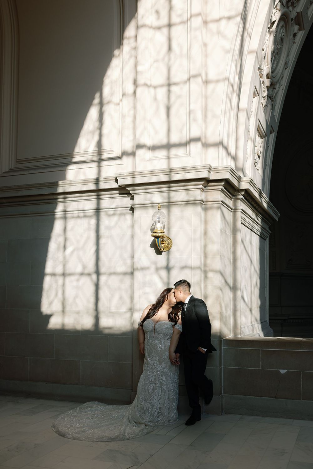A couple in formal wedding attire stand against a white stone wall with dramatic window shadows creating an architectural pattern.
