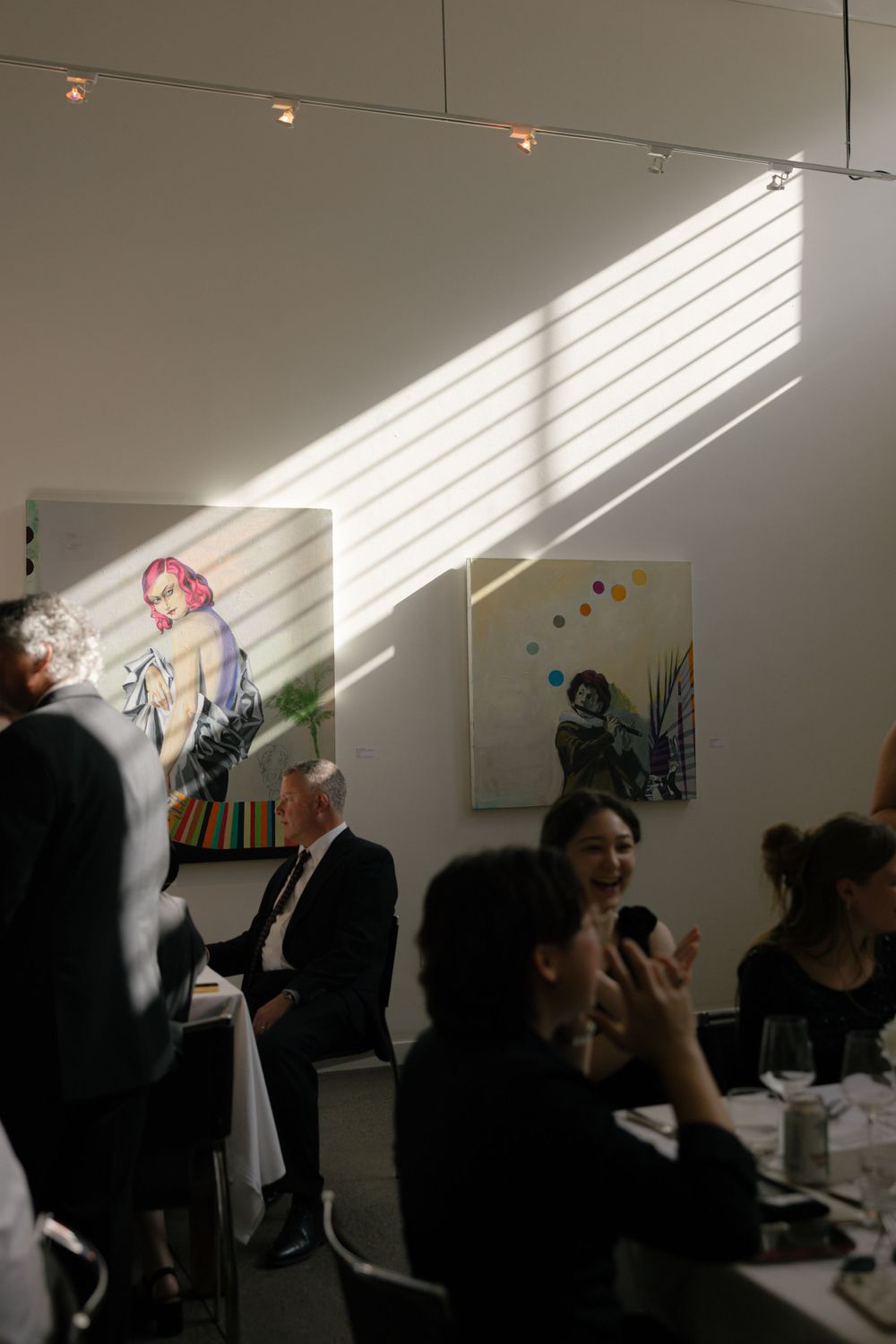 Light streams through windows in an art gallery as people gather at tables beneath colorful artwork on display.