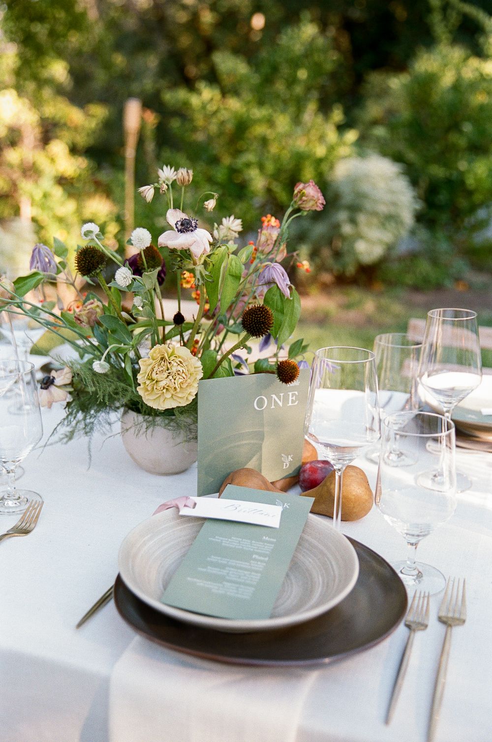 Elegant outdoor table setting with soft floral centerpiece, white linens, charger plates and menu cards in sage green tones.