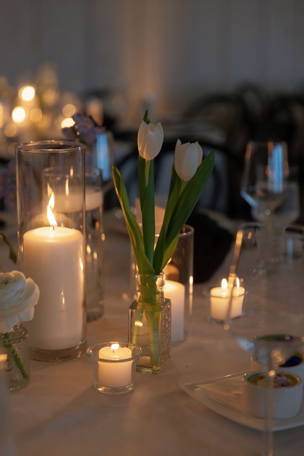 White tulips in glass vase alongside glowing candles and table settings create an elegant dinner ambiance.