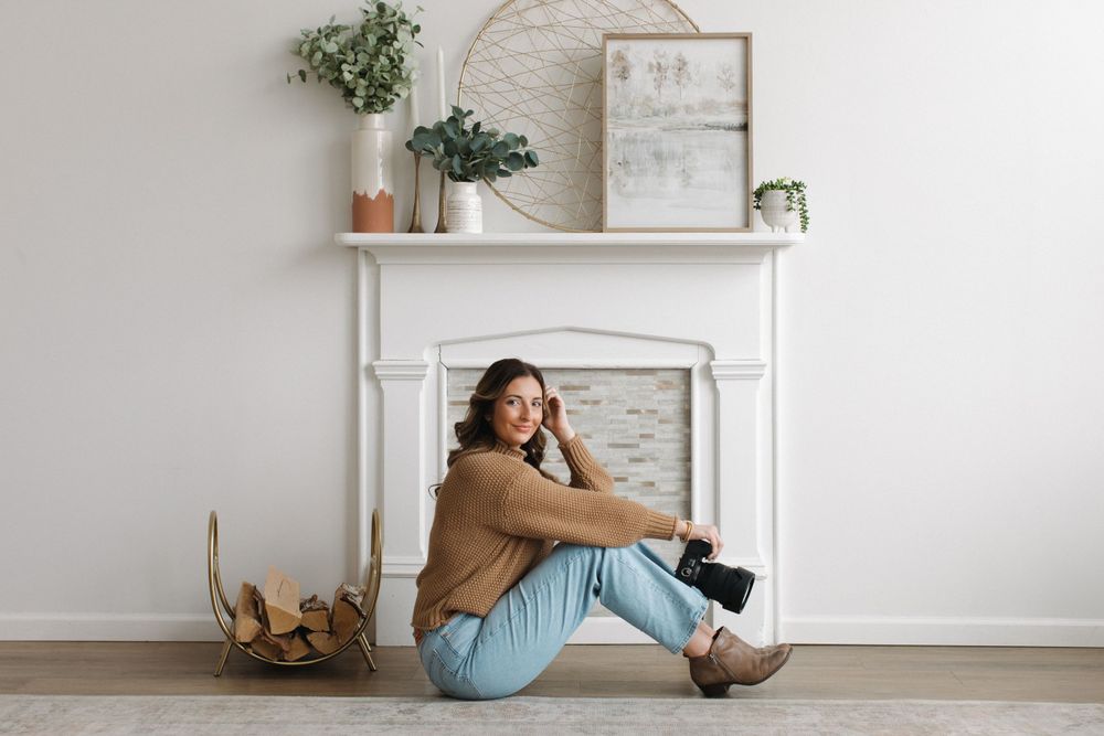 Casual photographer with brown boots sitting on floor by white fireplace with plants and decor on mantel.