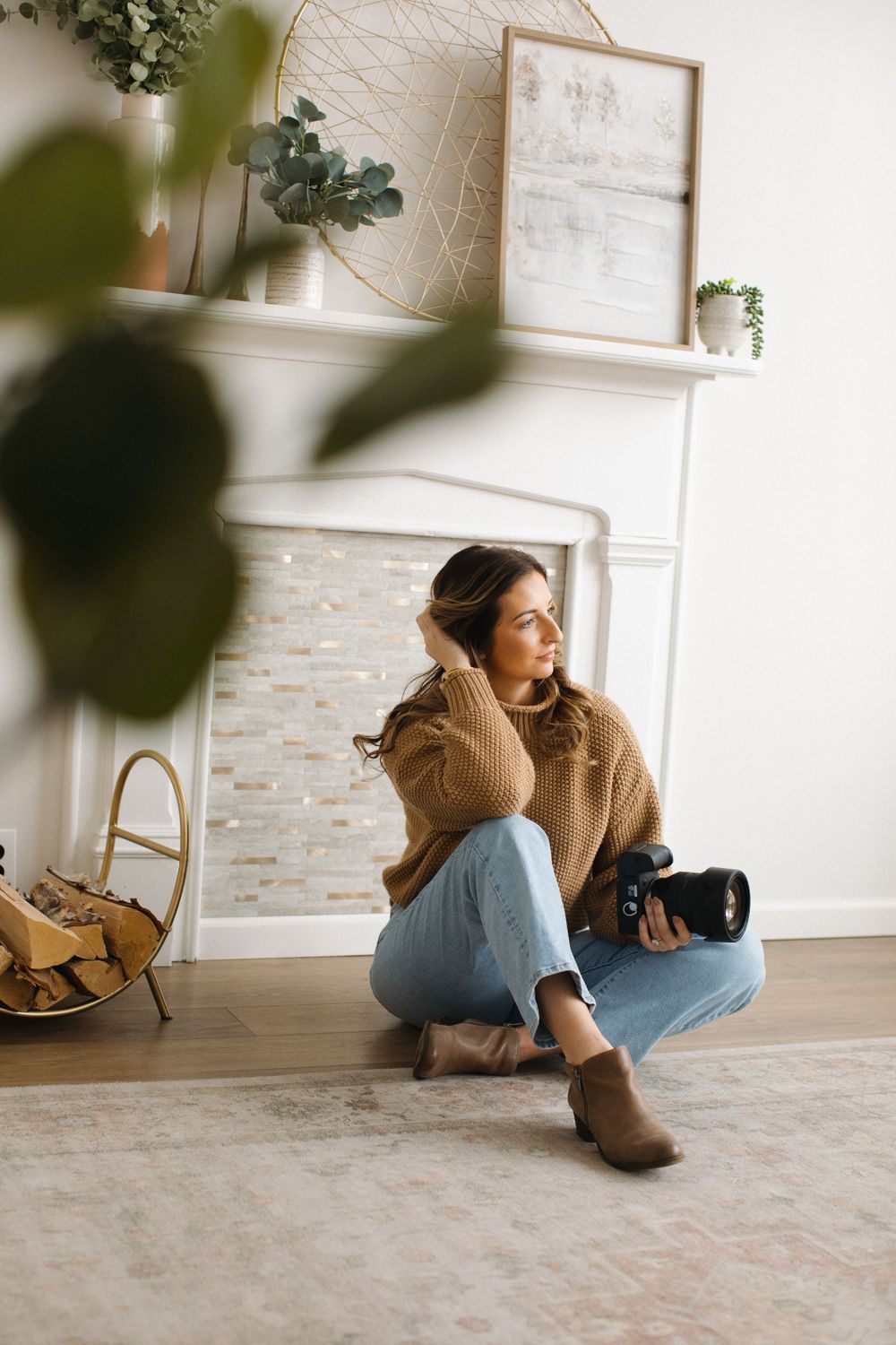 A photographer wearing jeans and brown sweater sits cross-legged on floor near fireplace with camera and home decor.