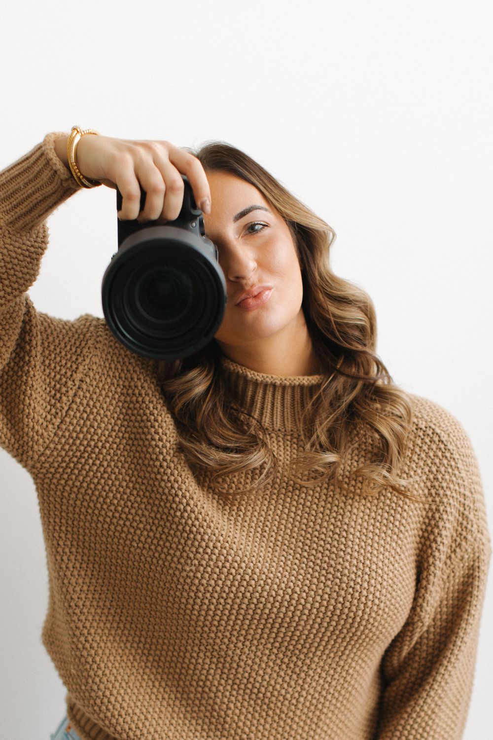Photographer in a cozy brown knit sweater holds up a professional camera while standing against white background.
