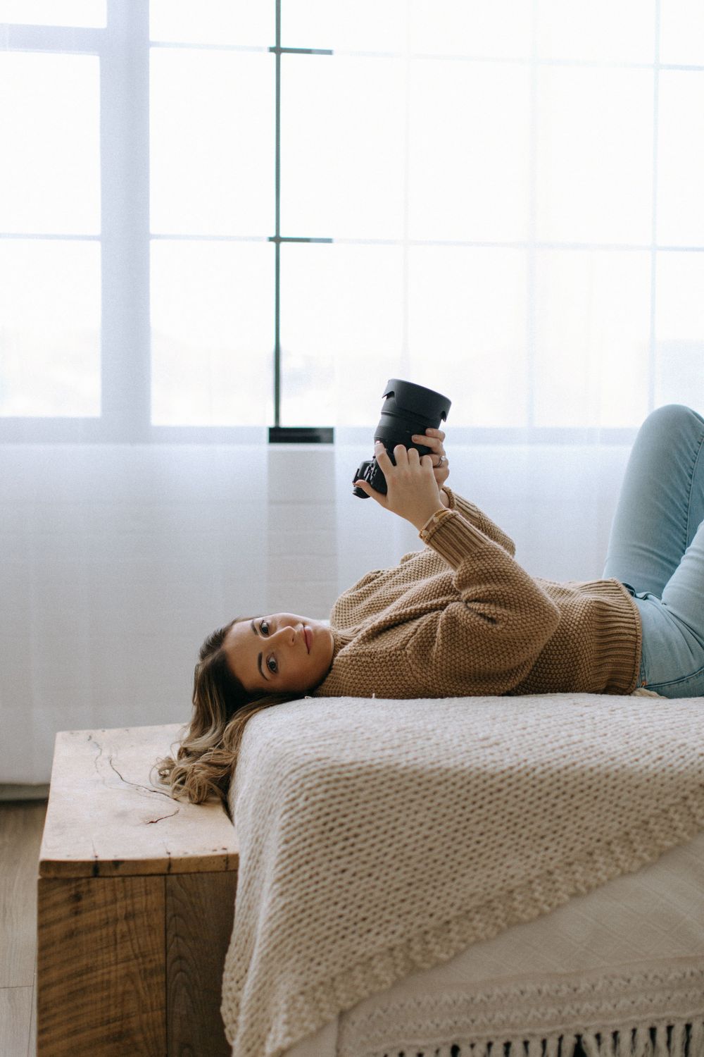 Relaxed woman in cozy sweater lies on bed holding coffee mug while looking at camera near window.