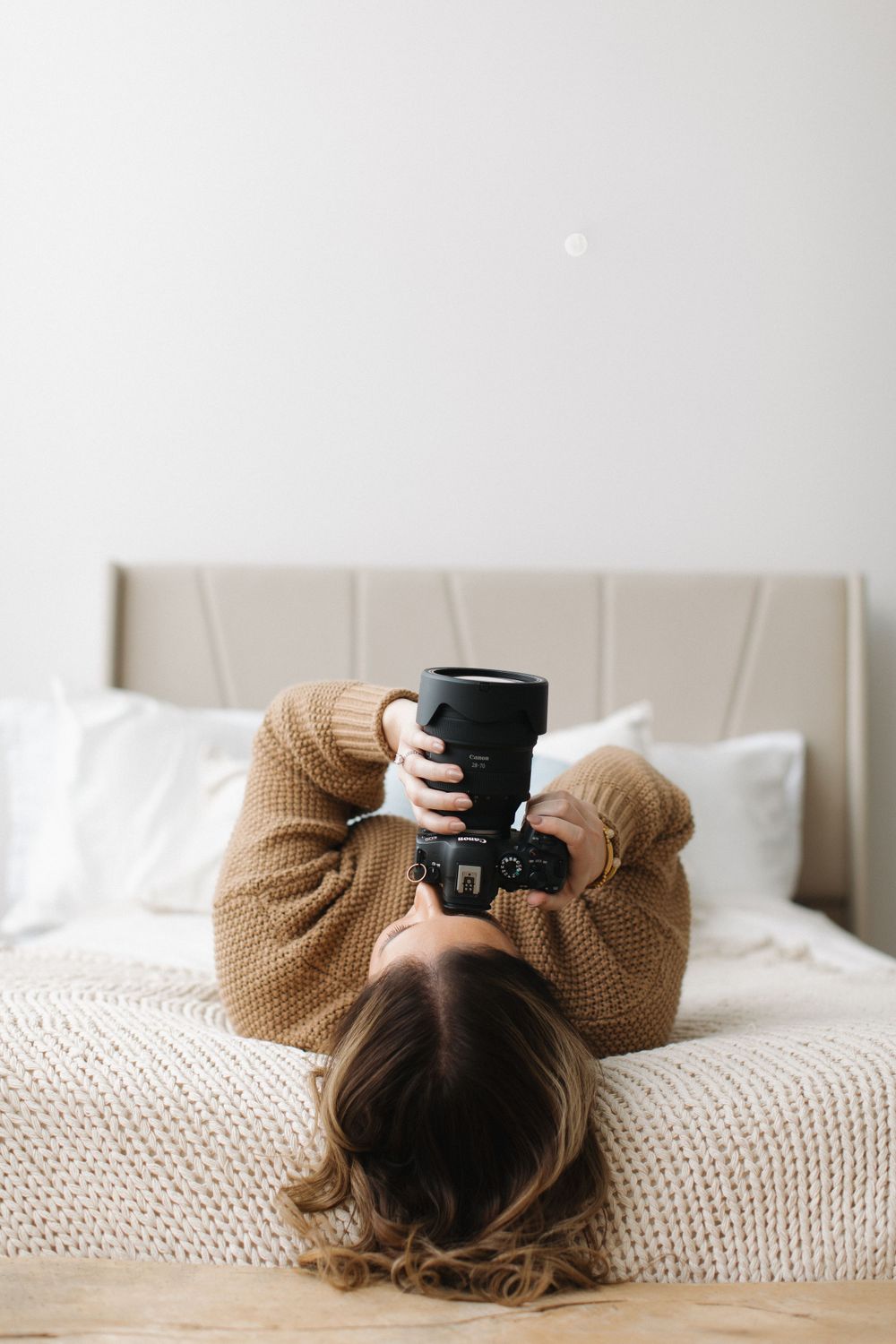 Person lying on white bedding wearing cozy brown sweater while holding black coffee mug in relaxed position.