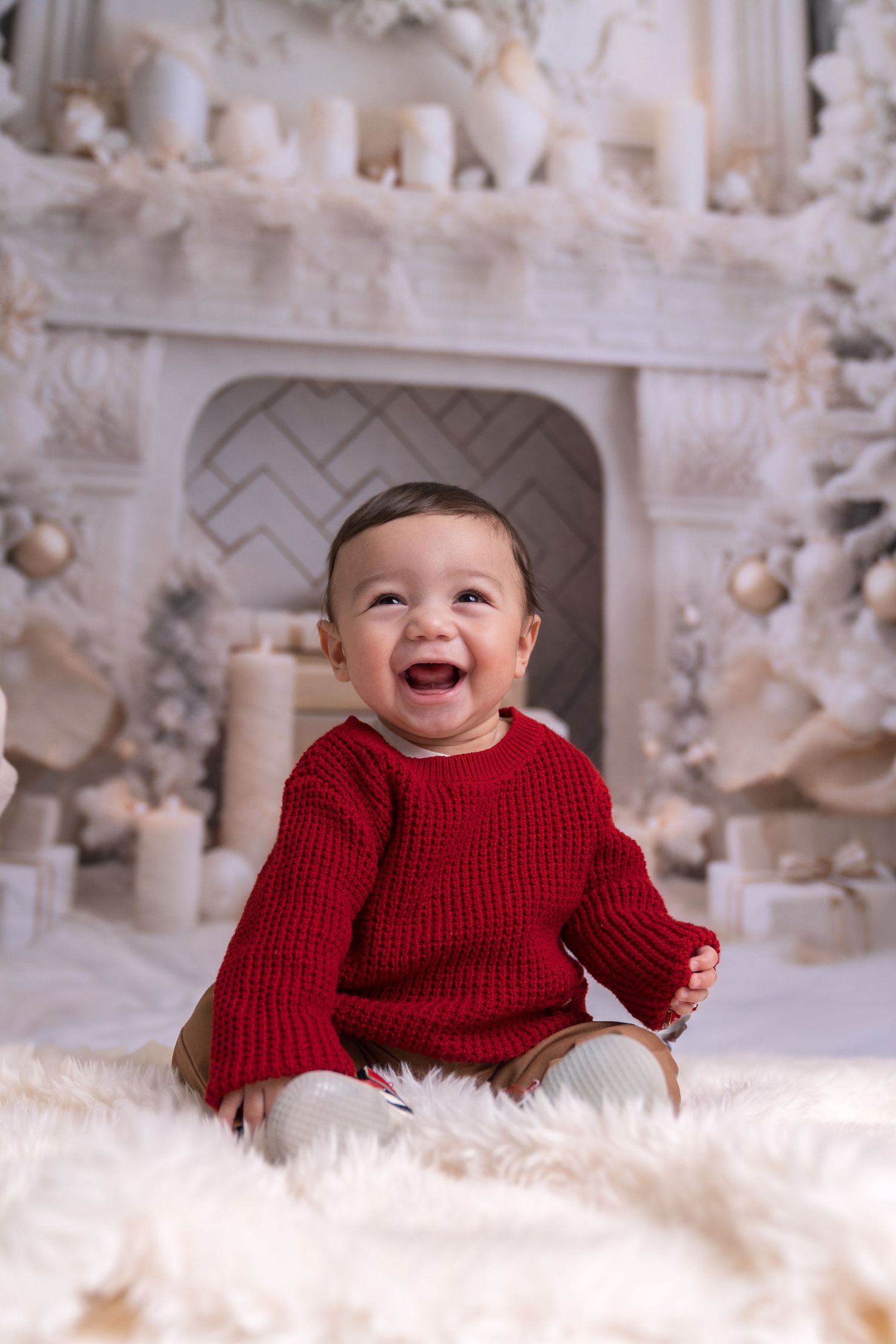 Baby in red sweater smiling while lying on white fur with Christmas decorations in background.