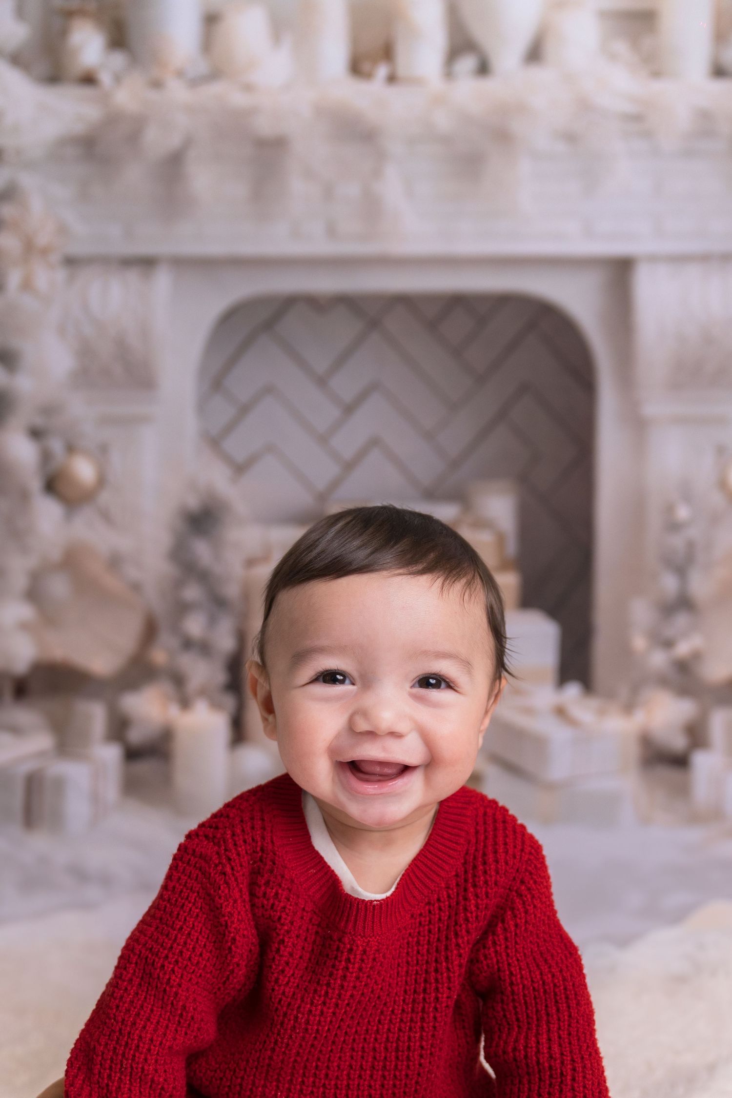 Happy baby wearing red knit sweater poses against white holiday backdrop with fireplace.