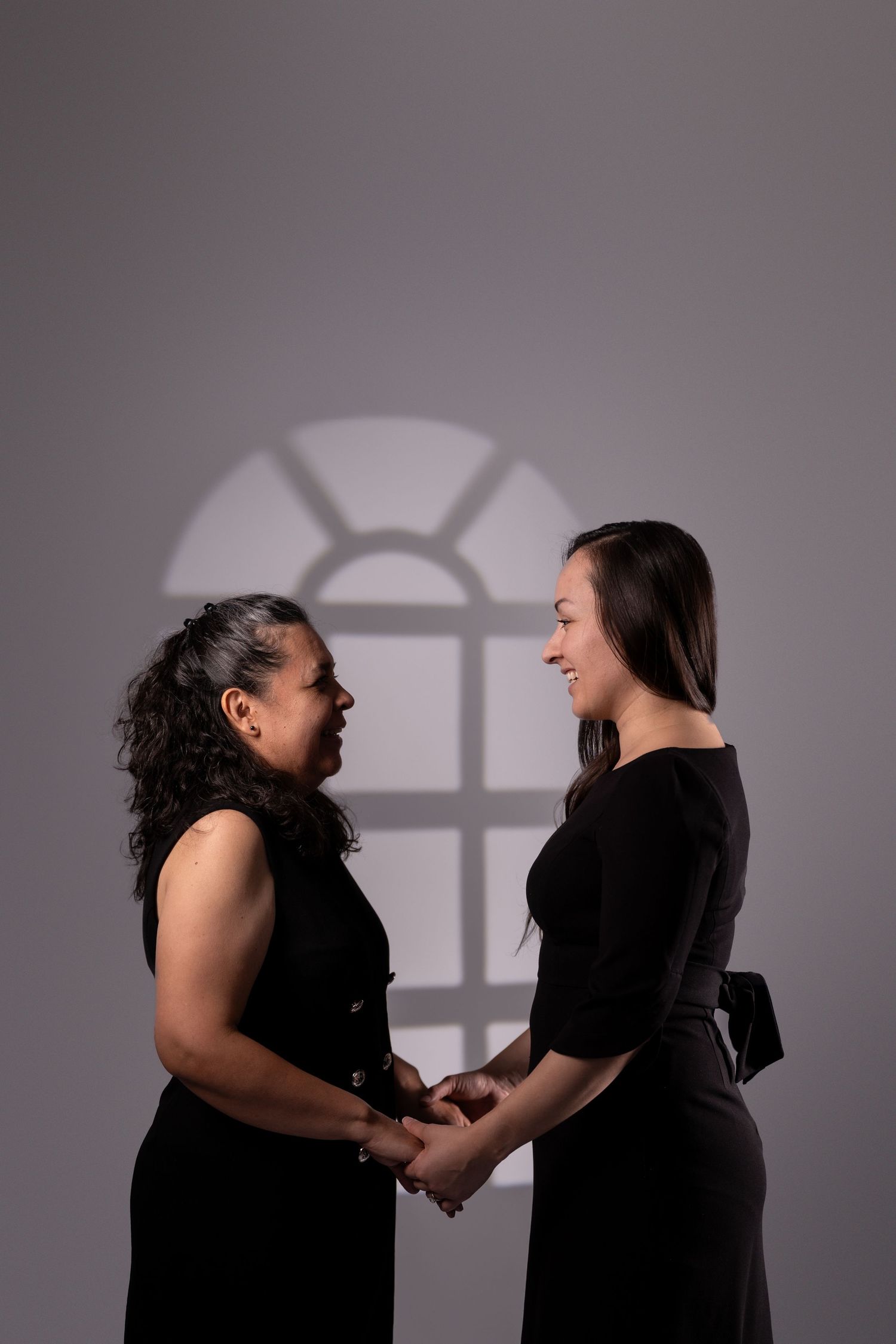 Two people in black dresses hold hands while standing in front of a window shadow projection.