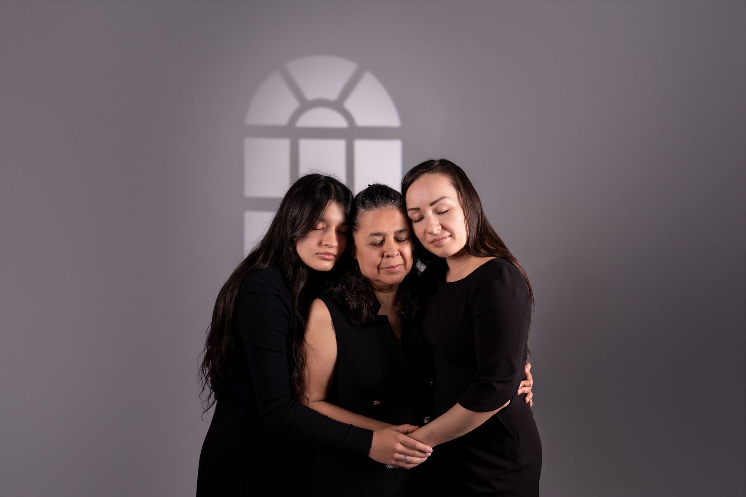 Three people in black outfits embrace and laugh together under a window shadow.