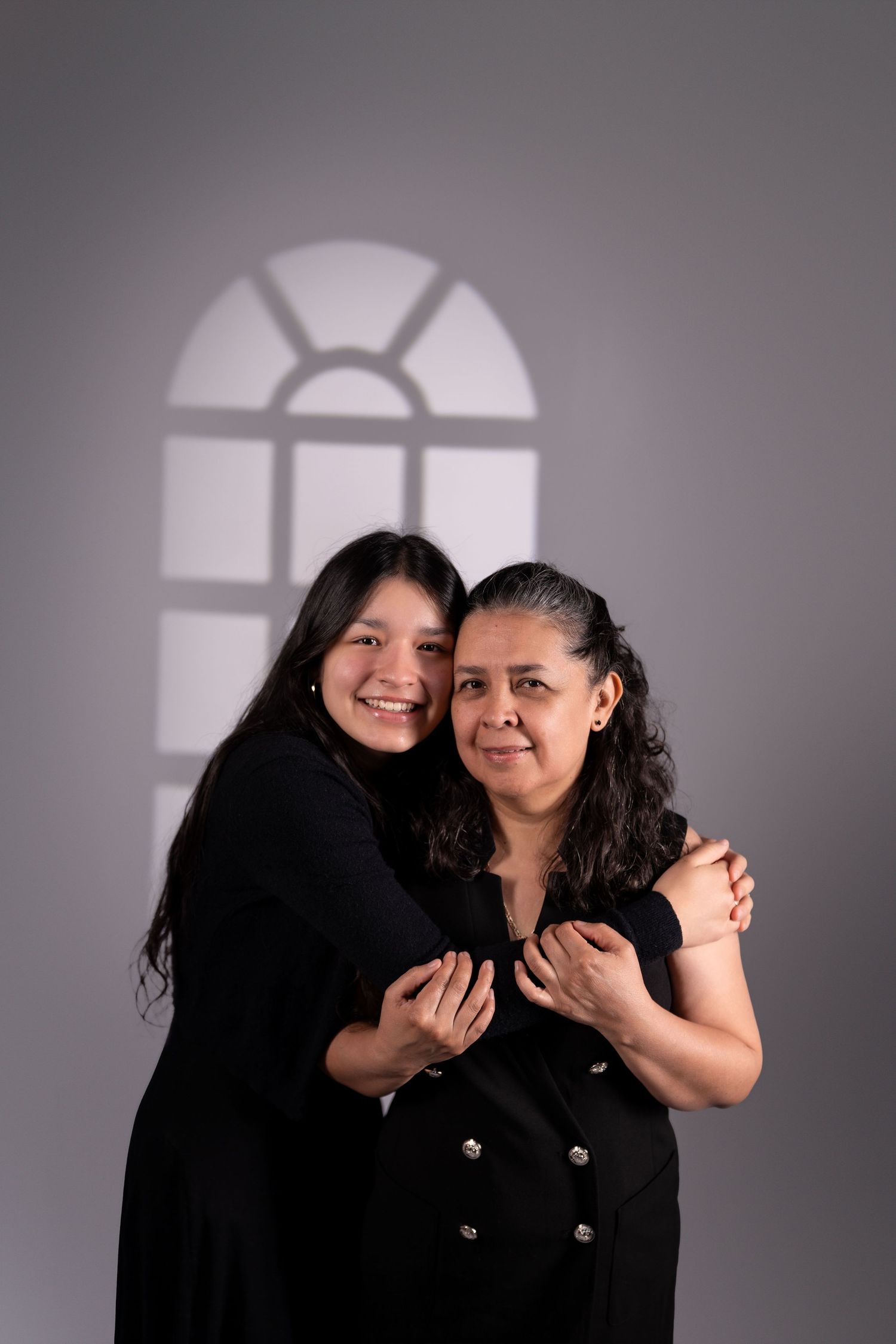 Two people in black dresses share a warm embrace under an arched window shadow.