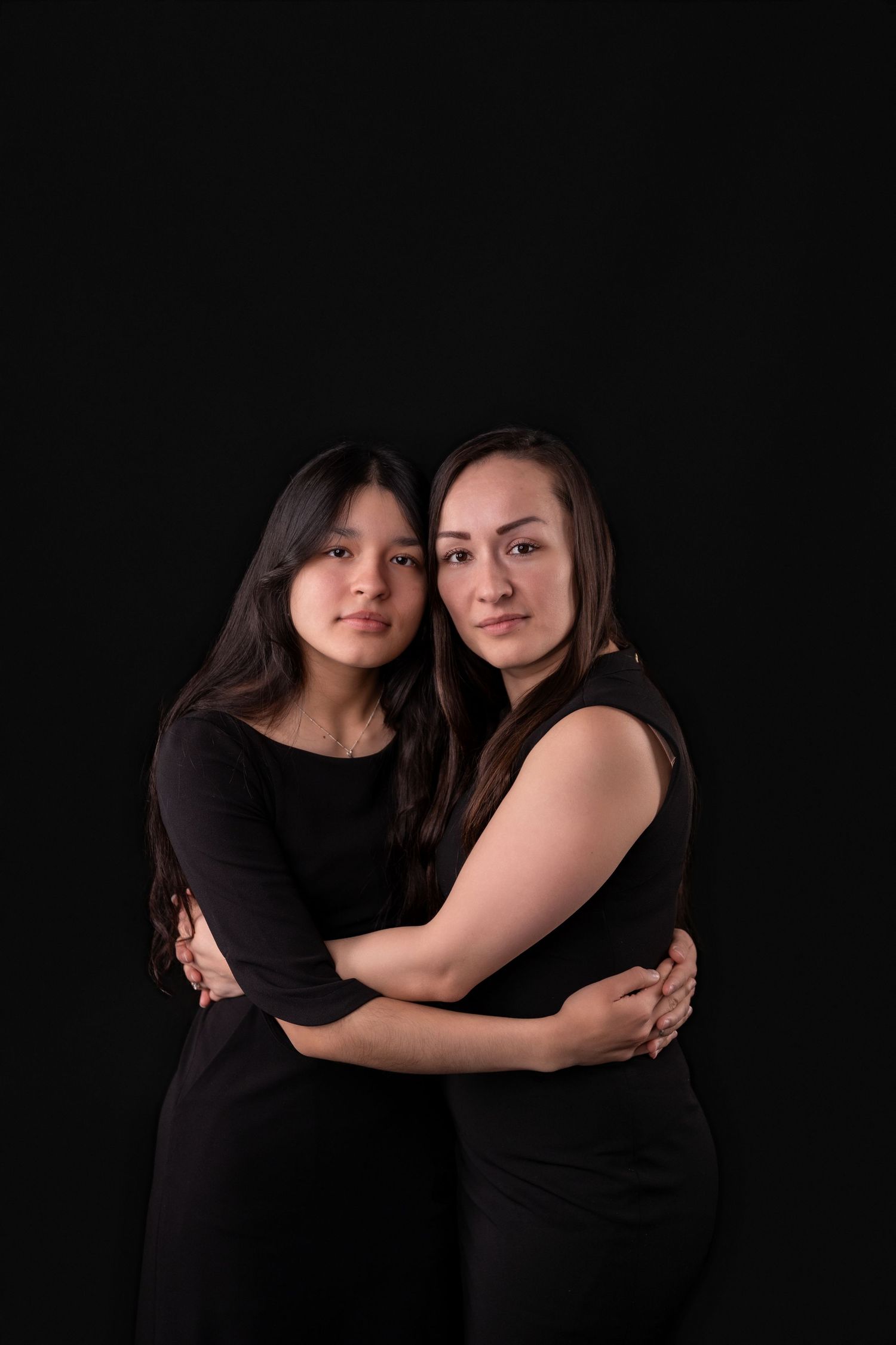 Two people in black outfits embrace against a dark background in a studio portrait.