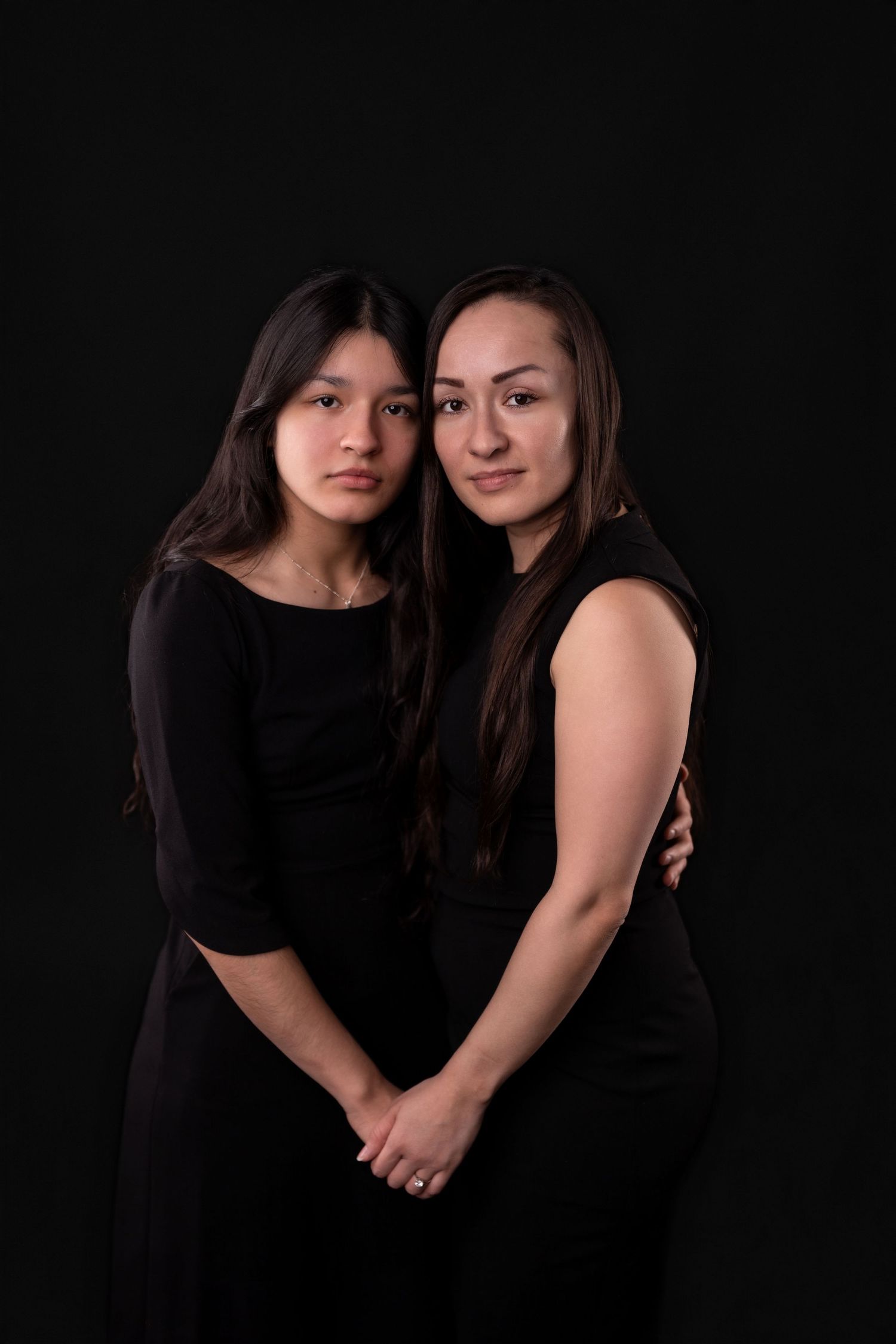 Two people in black dresses pose together for an intimate portrait against dark background.