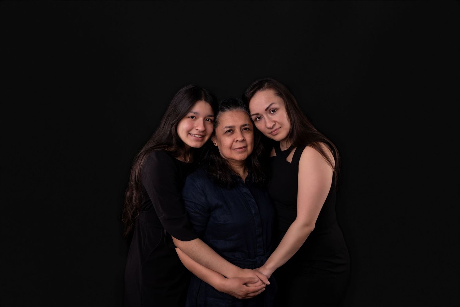 Three people in dark clothing embrace in a loving group portrait.