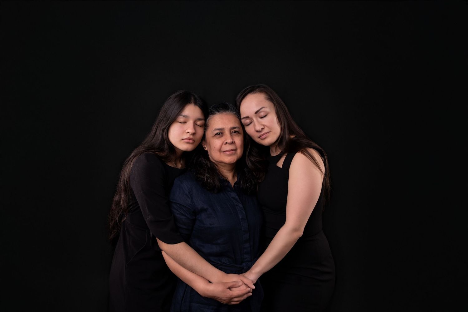 Three people in dark outfits share a warm family embrace against black background.