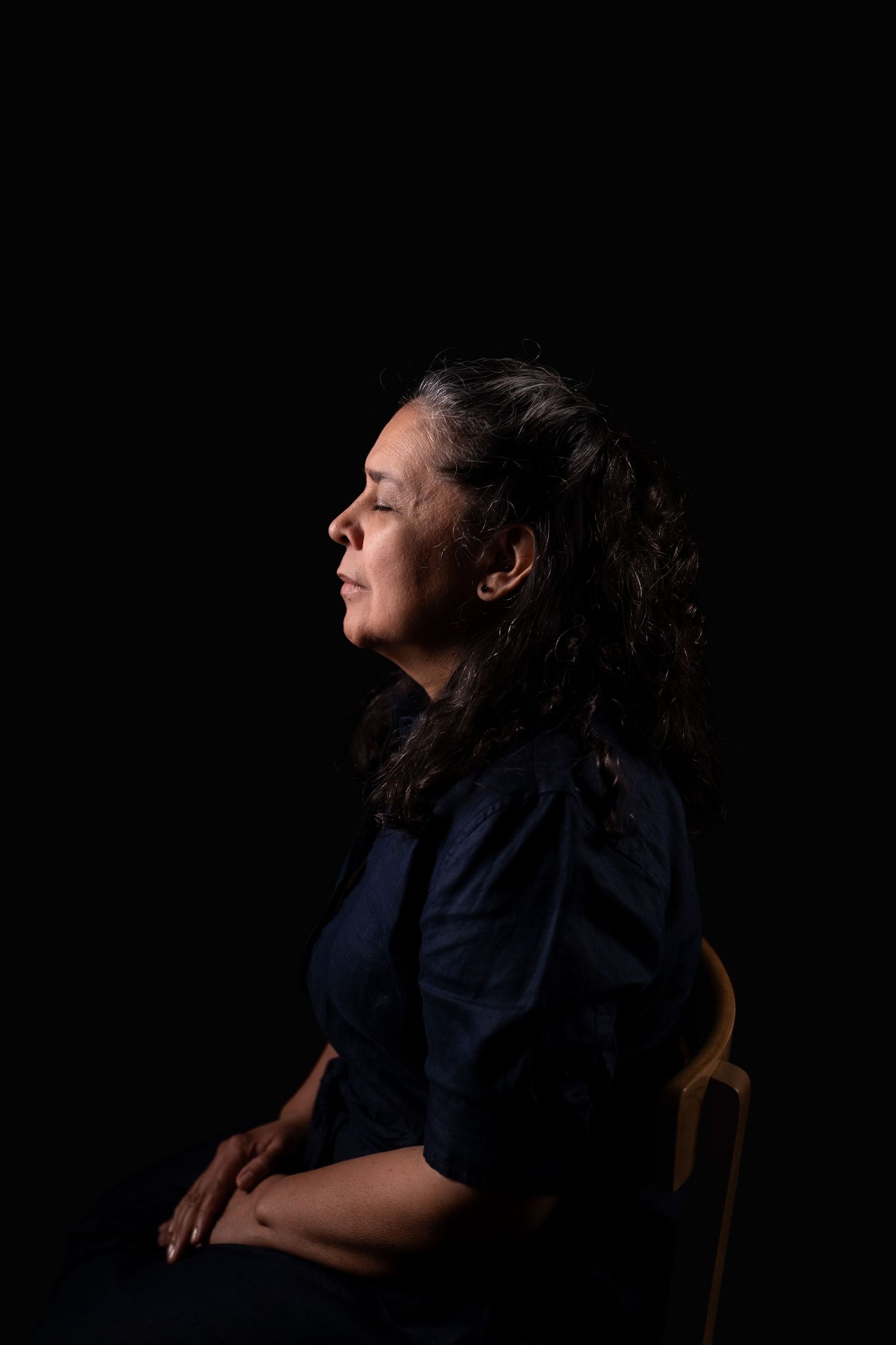 Person with long dark hair sits in profile against black background in dramatic lighting.
