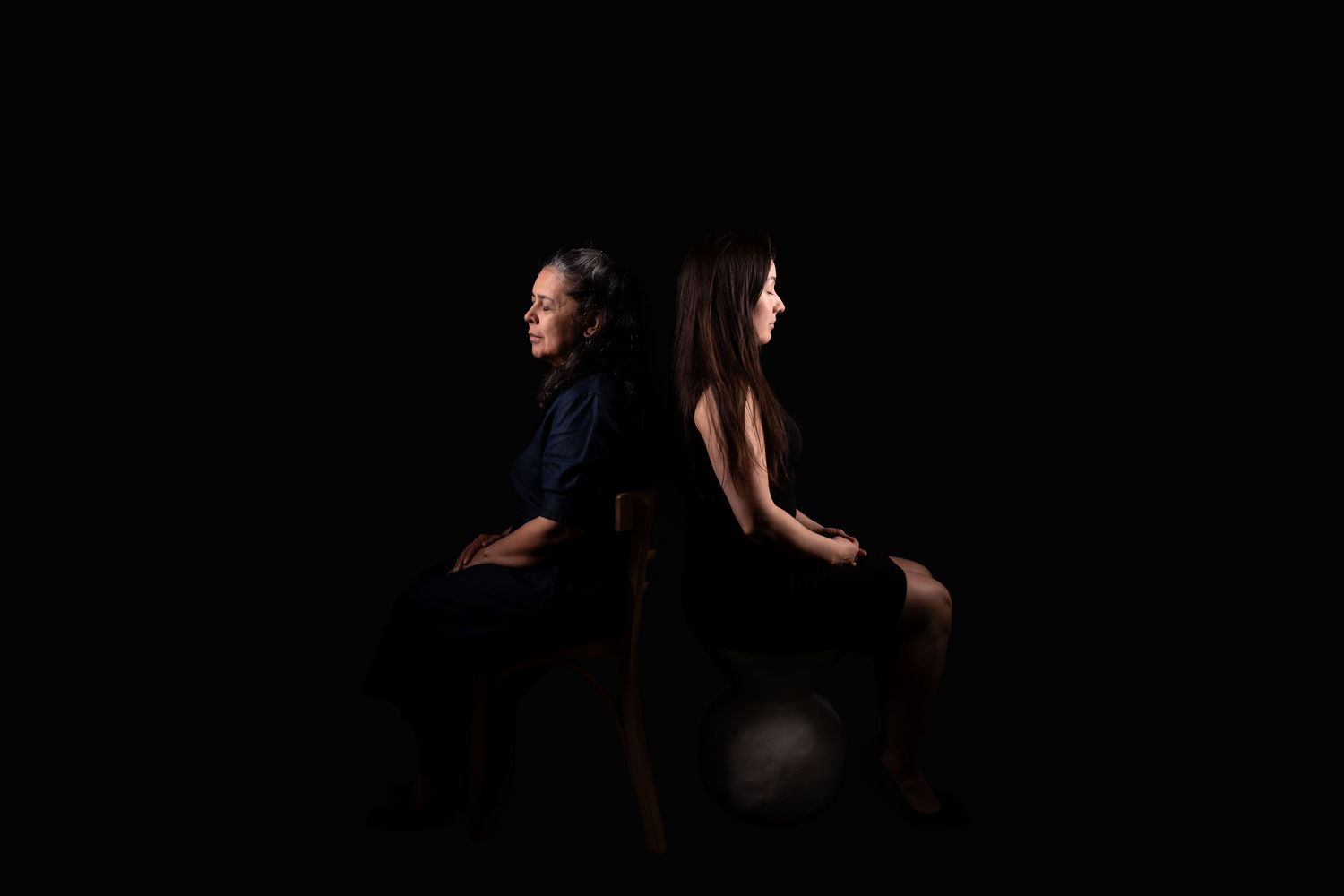 Two people in dark clothing sit back to back in dramatic studio lighting.
