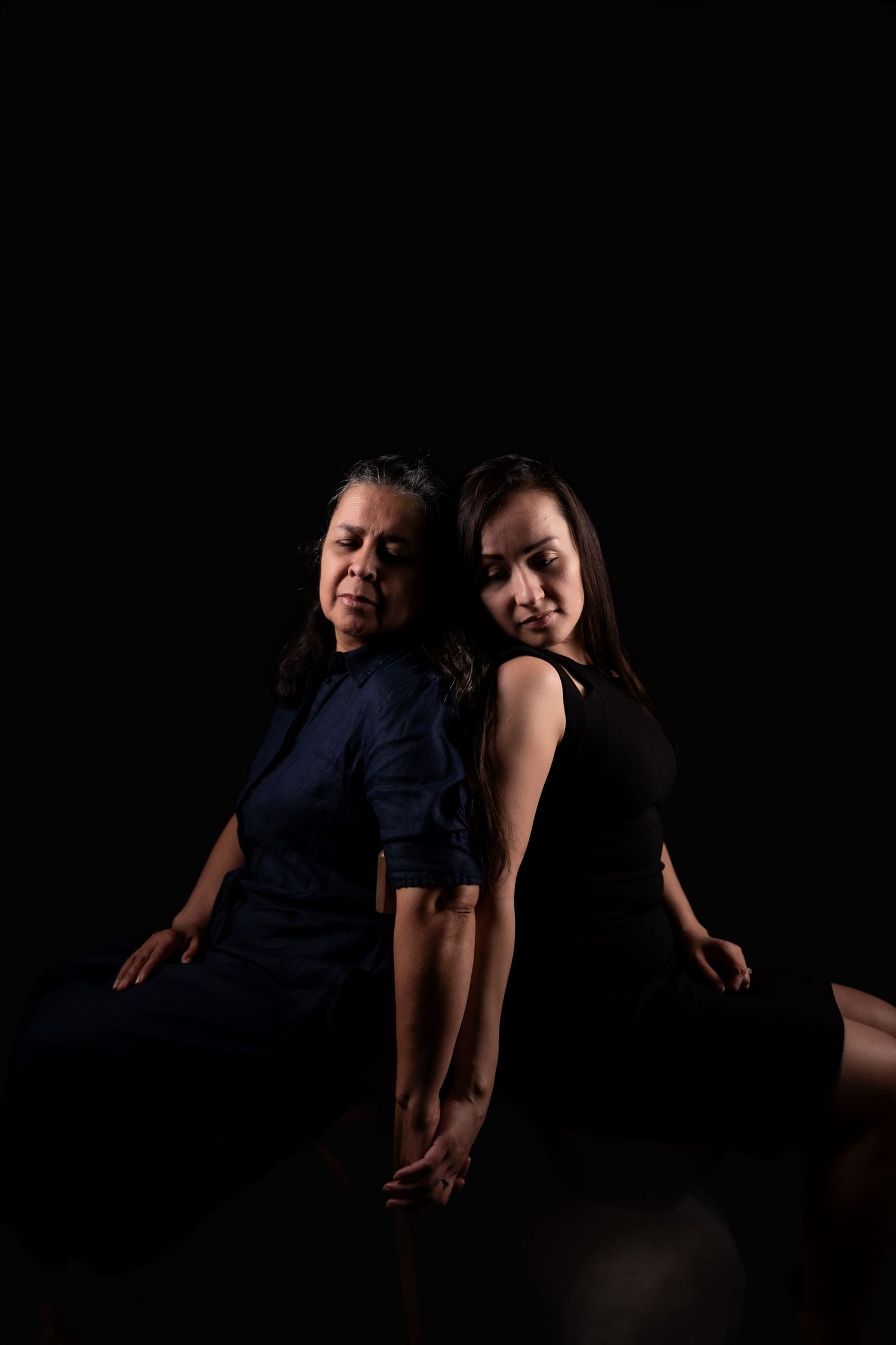 Two figures embrace in a moody black and white portrait against dark backdrop.
