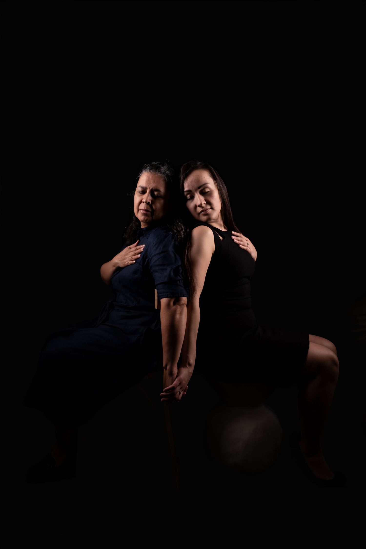 Two people pose together against a dark background in dramatic low-key lighting.