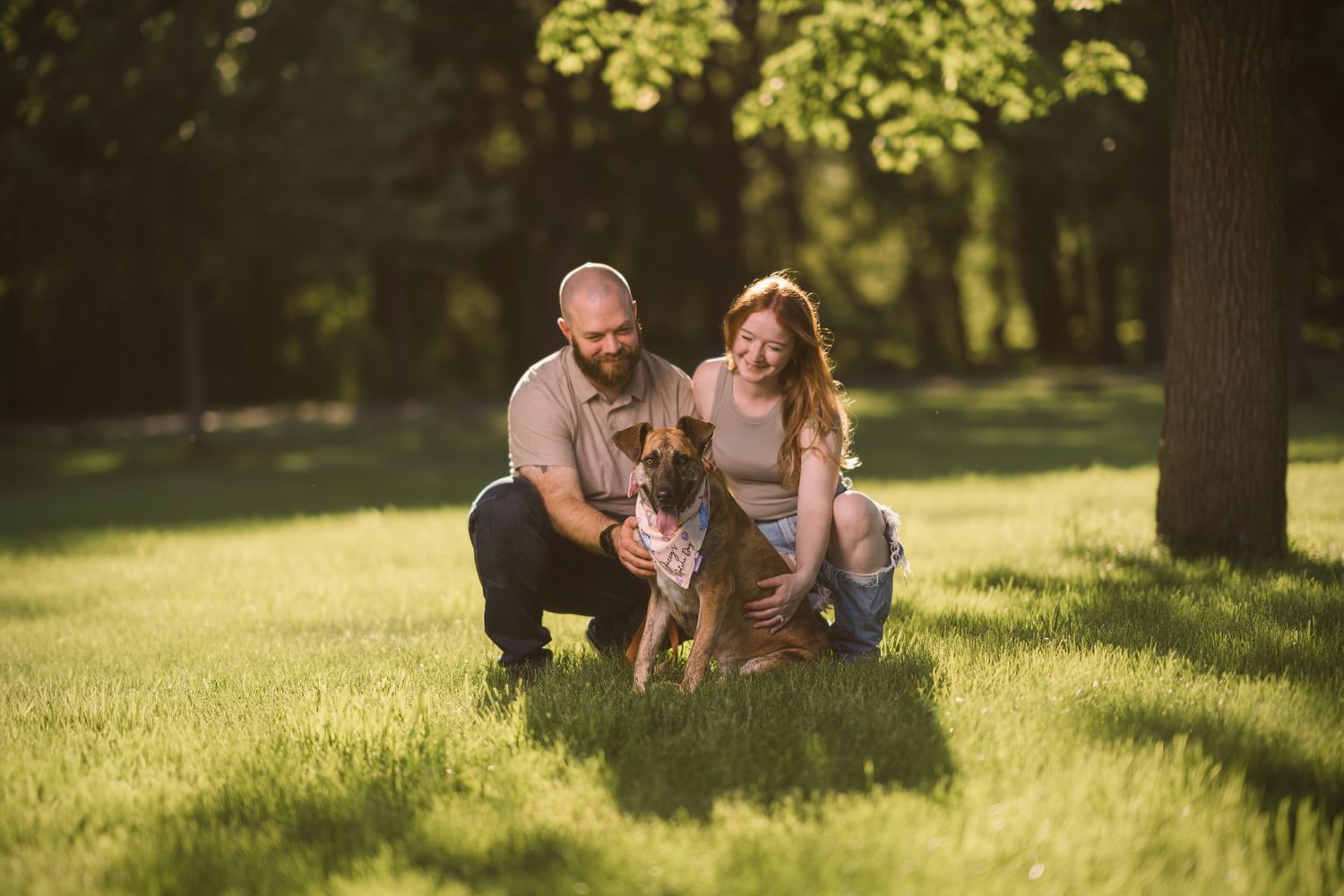 A couple and their dog enjoy a sunny afternoon in a grassy park setting with golden sunlight filtering through trees.