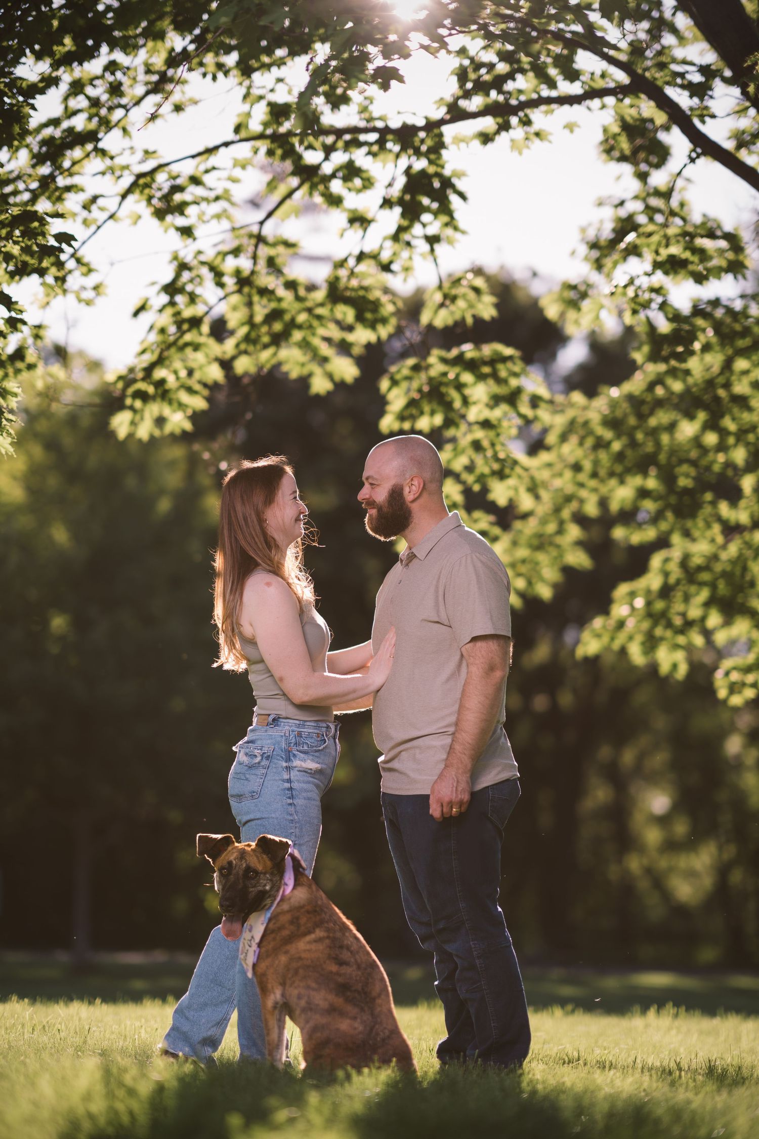 A couple stands together in a park at sunset with their loyal dog sitting beside them.