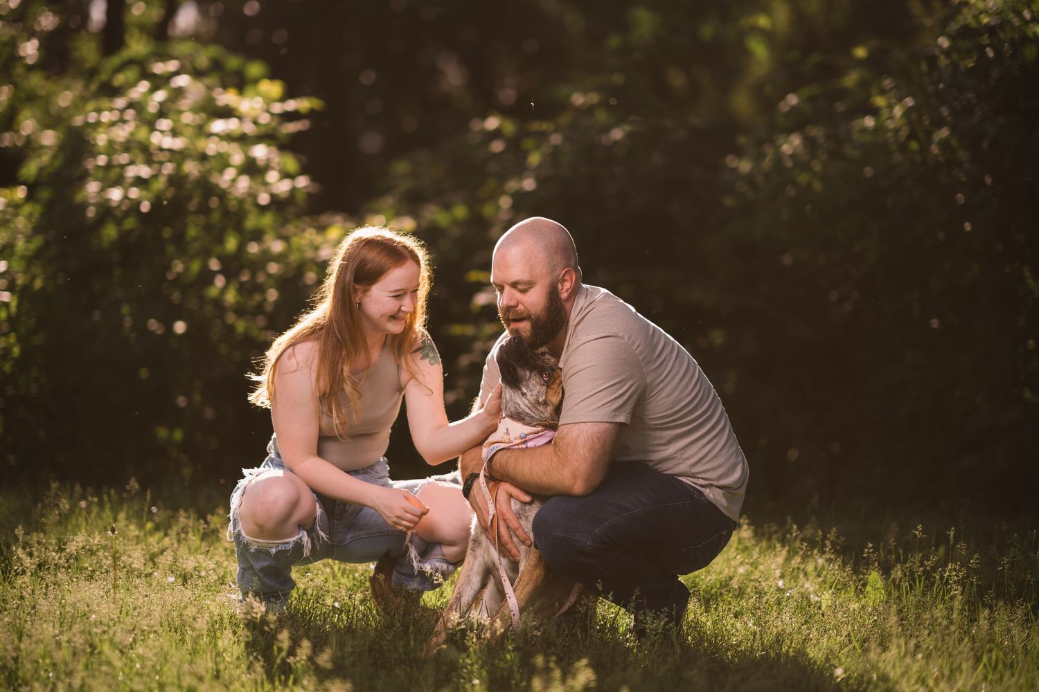 A couple crouches down to play with their dog in a sunlit garden setting.