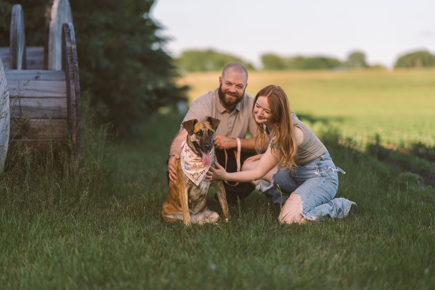 A couple poses with their brown dog near a rustic fence during sunset.
