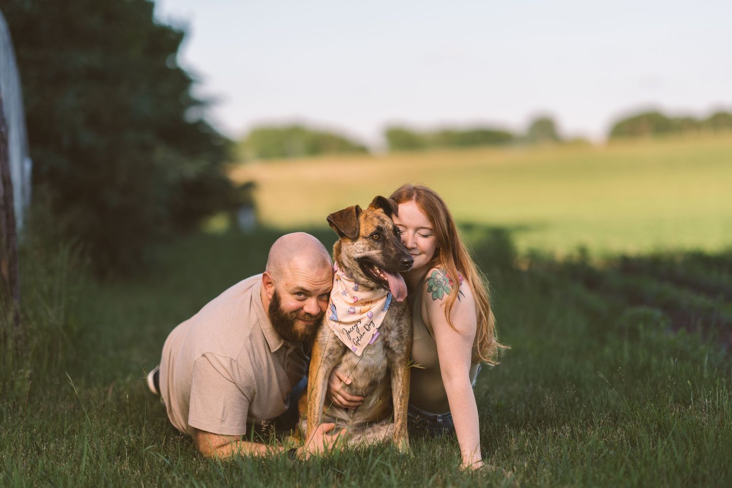A couple poses with their friendly dog in a serene field at dusk.