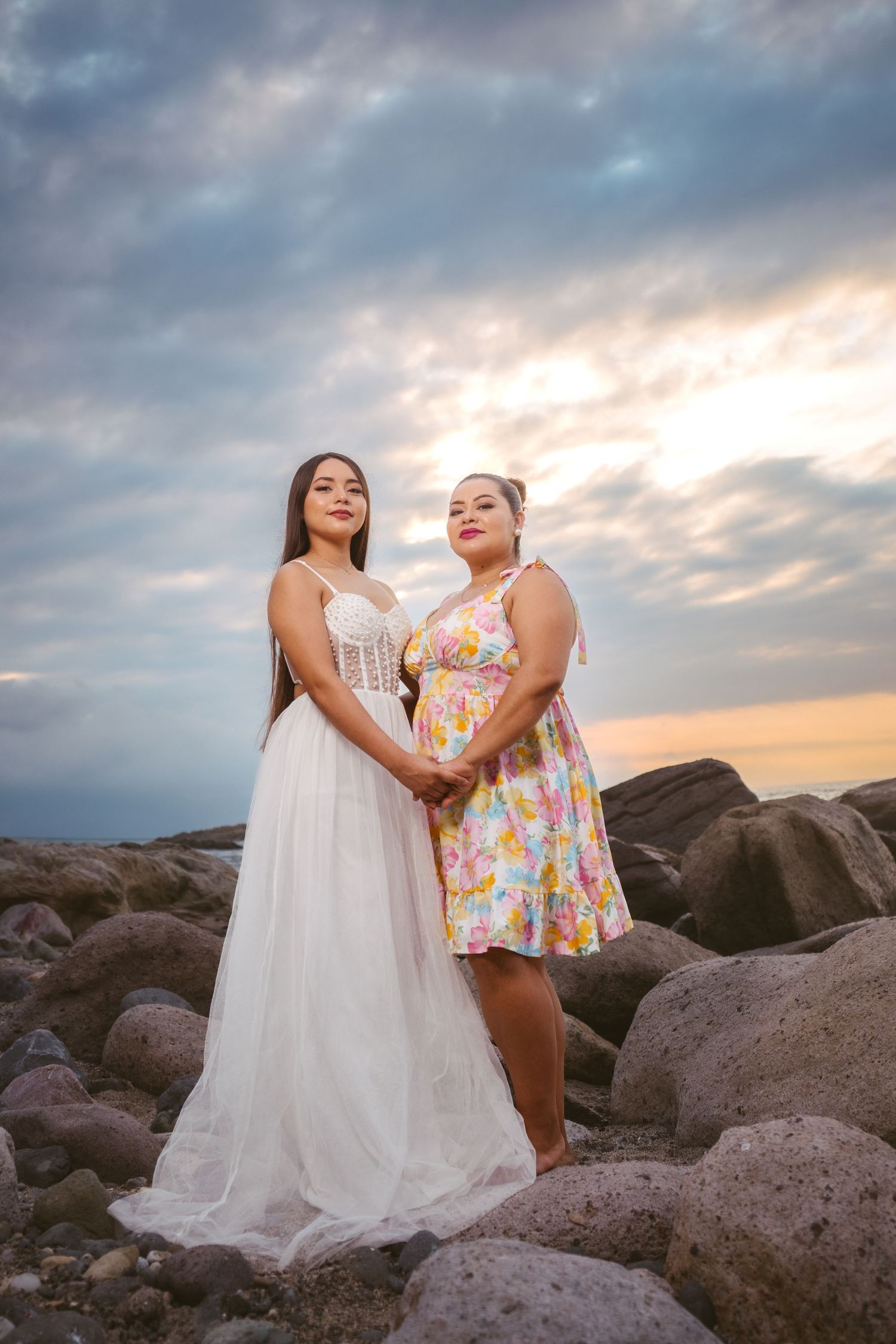 Two people in white and floral dresses pose together on rocky coastal terrain at sunset.