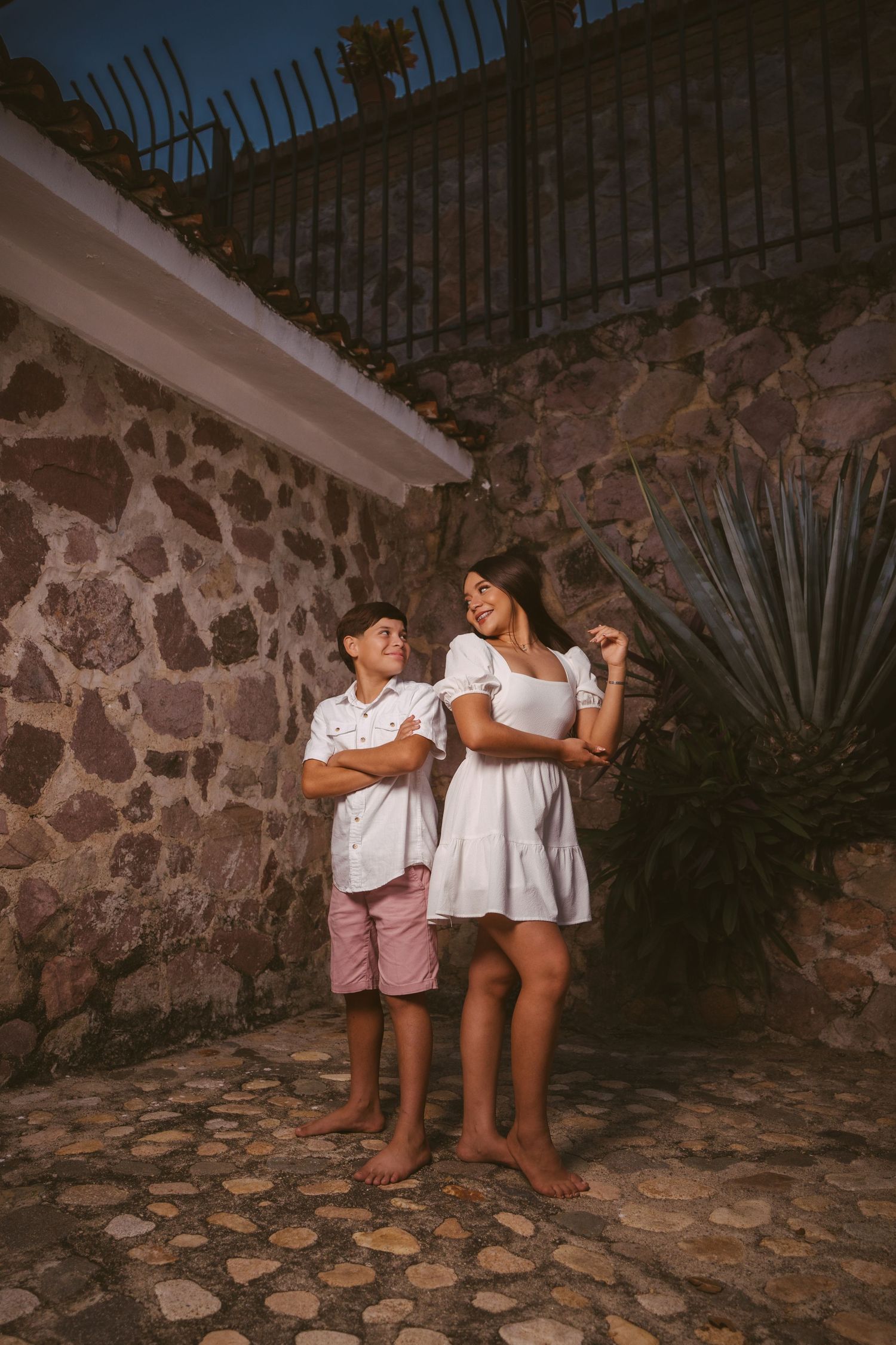 Siblings share candid moments against a textured stone wall backdrop at sunset.