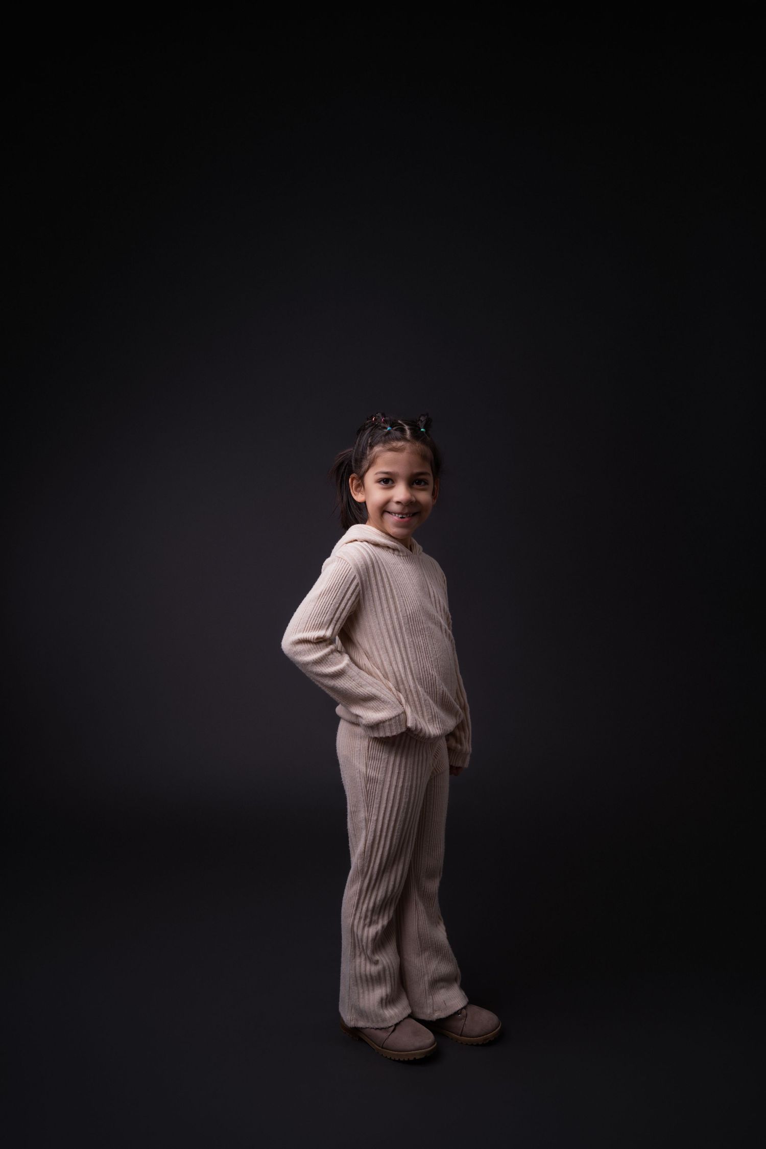 Child in beige knit outfit poses playfully against dark background.