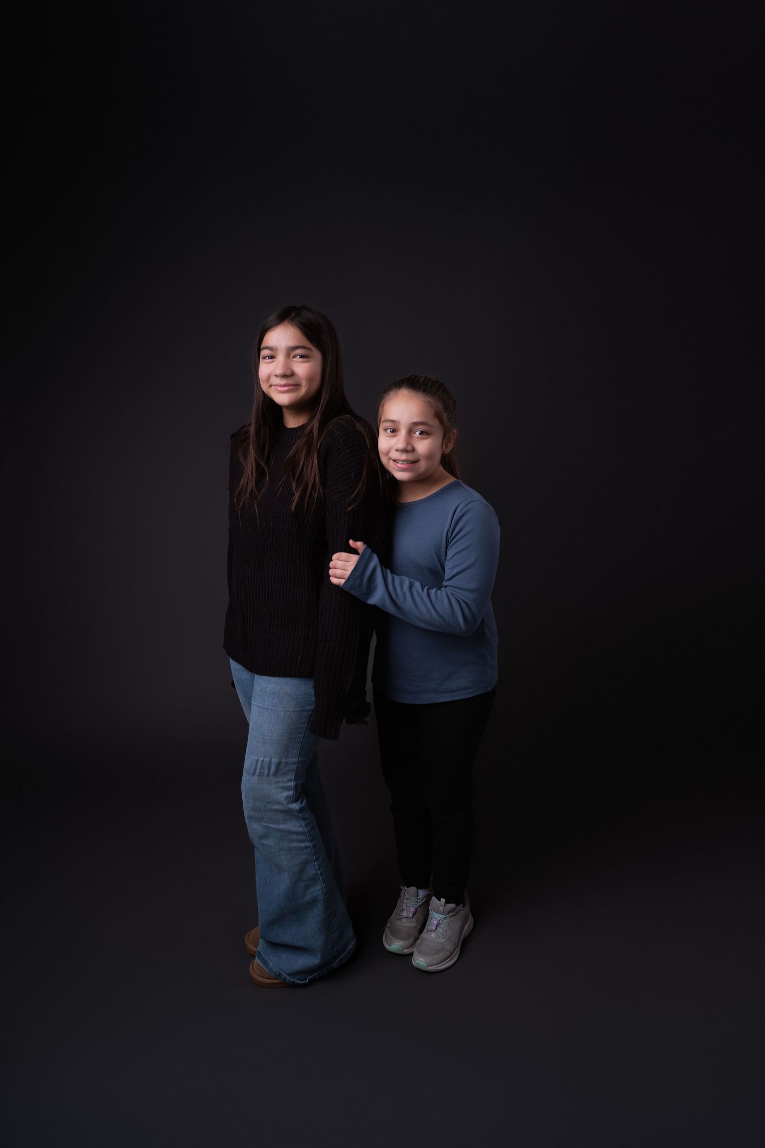 Two people in casual attire pose together against a dark background in a studio setting.