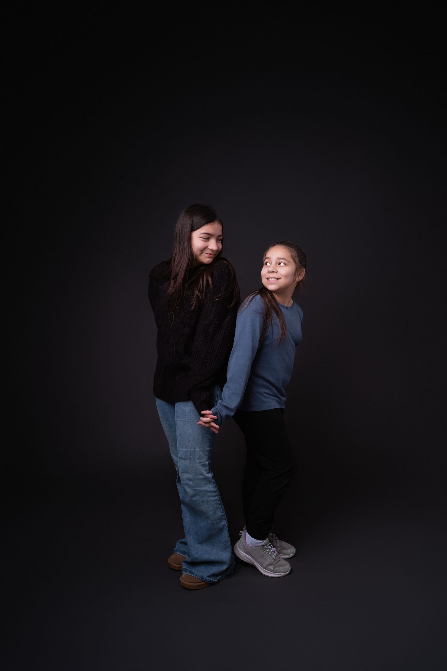 Two people in casual outfits crouch together during studio photoshoot with dark background.