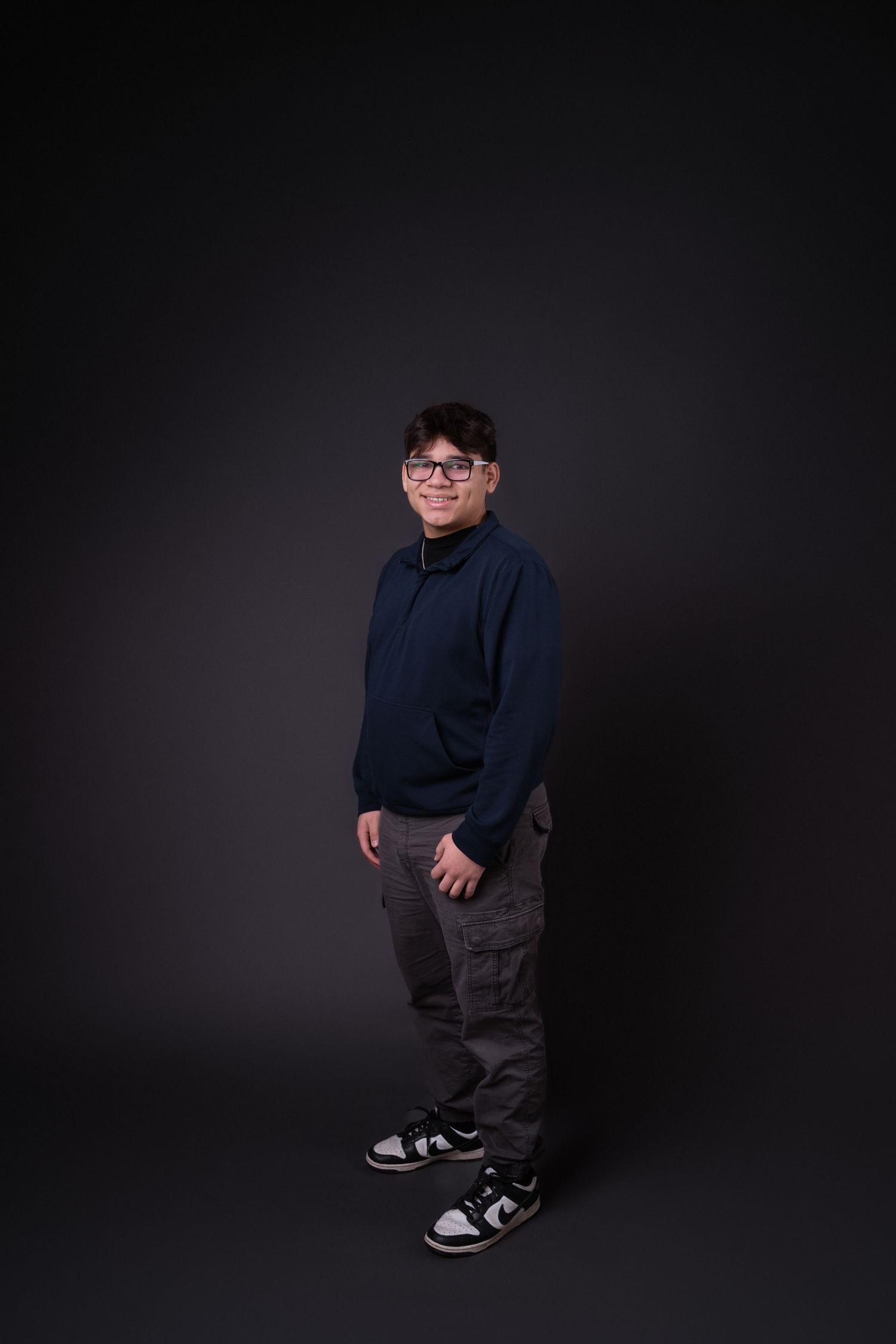 Person in navy sweater and gray pants poses casually against dark backdrop.
