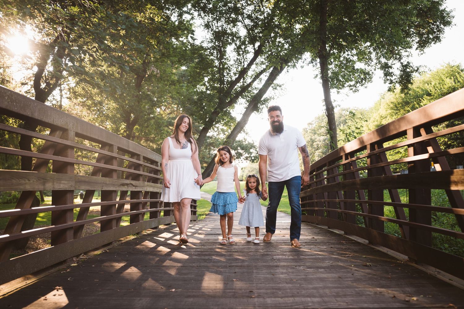 Family walking together on a wooden bridge path surrounded by lush trees during golden hour.