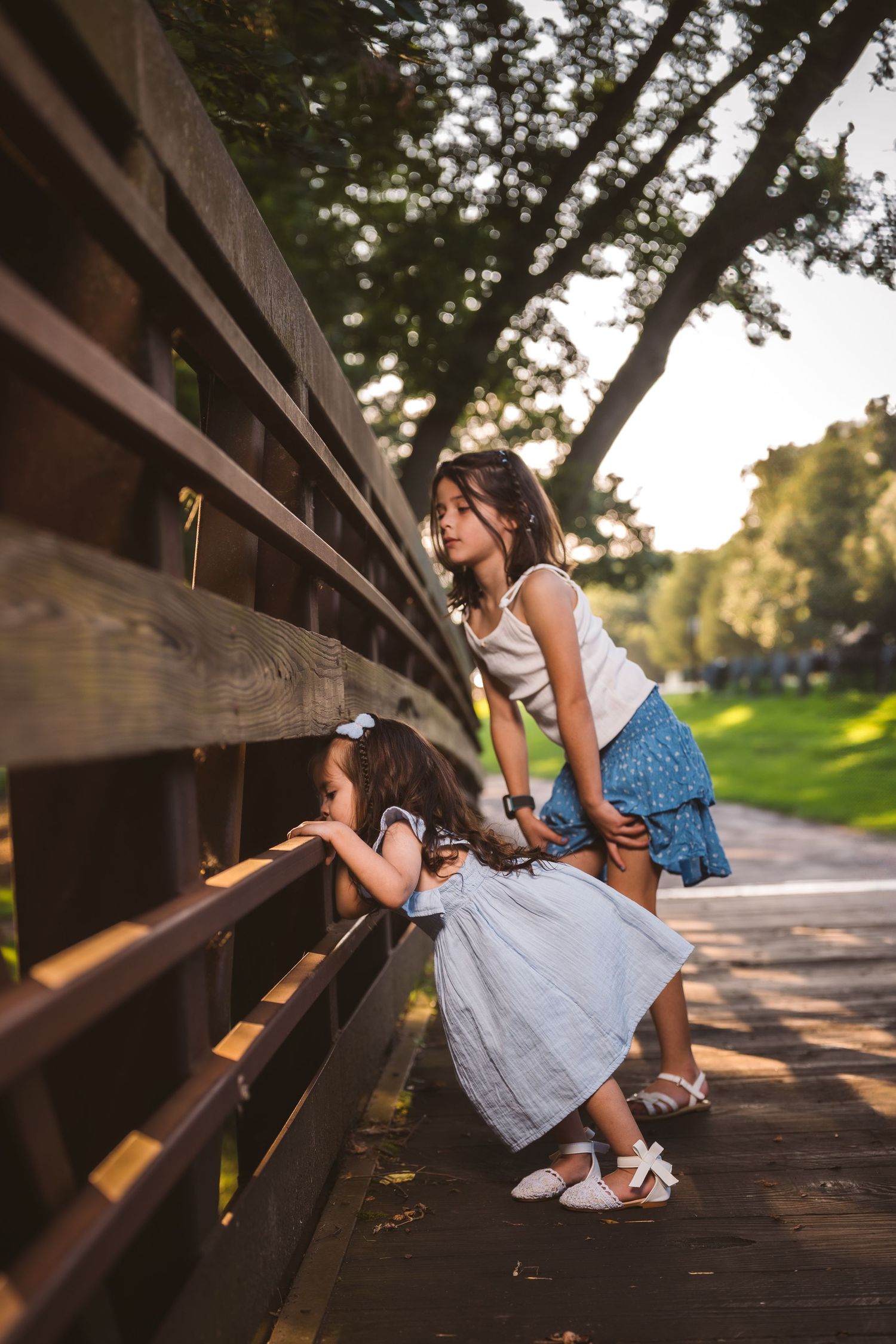 Young ones peek through wooden bridge railings during a sunny outdoor photoshoot.