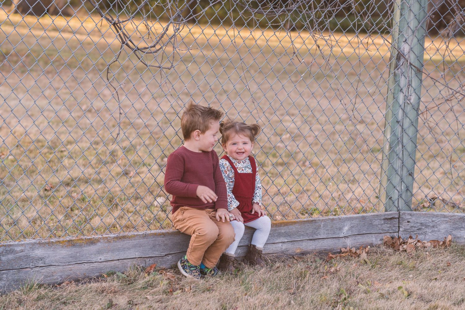Two children sitting together on wooden bench by wire fence in fall setting.