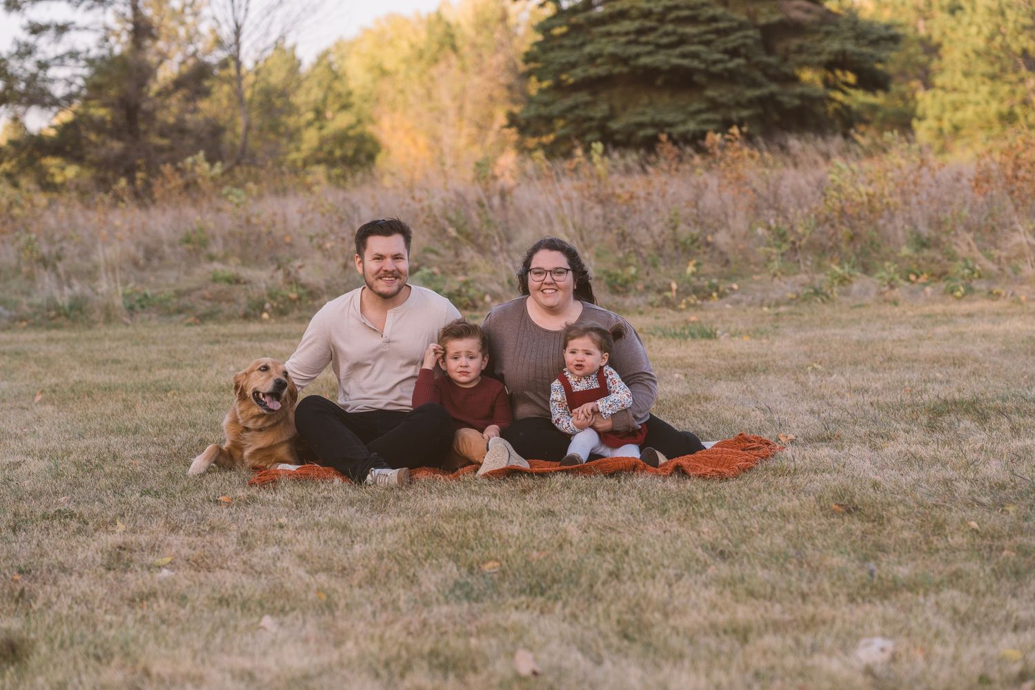 Family with dog relaxing on blanket in autumn field during sunset photoshoot.