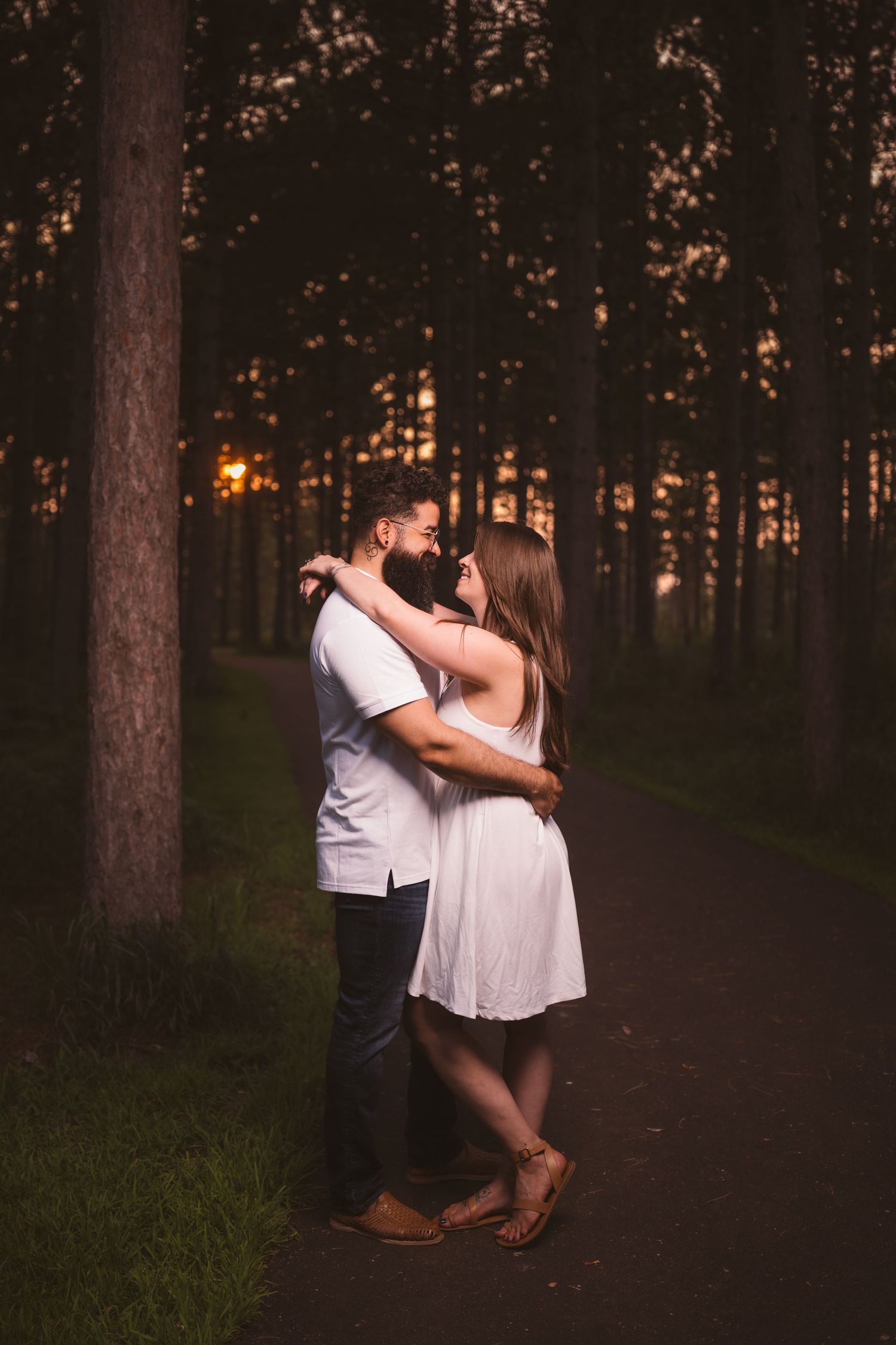 A romantic couple embraces on a wooded path during sunset, creating a silhouetted scene among tall pine trees.