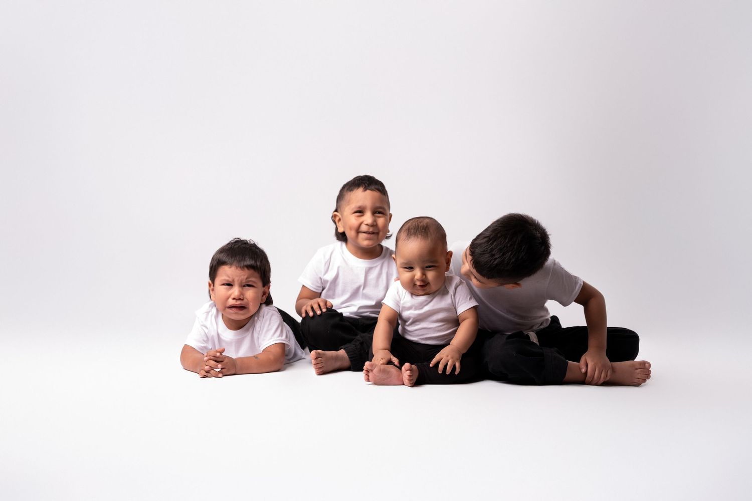 Four children lying on their stomachs in white shirts against a light background.