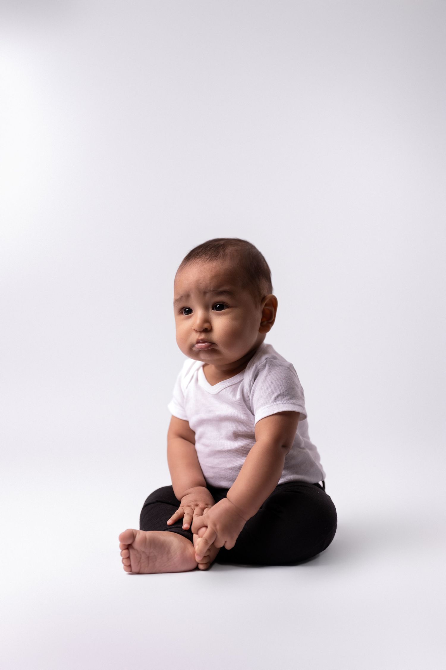 Baby wearing white shirt and black pants sitting on white background.