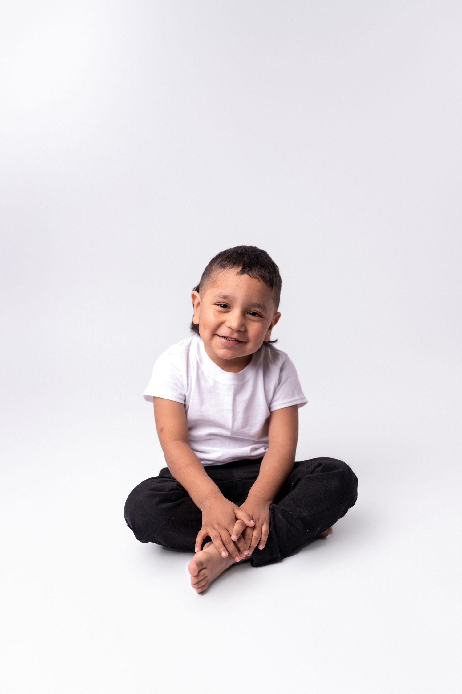 Young child in white t-shirt and black pants sitting cross-legged on white background.