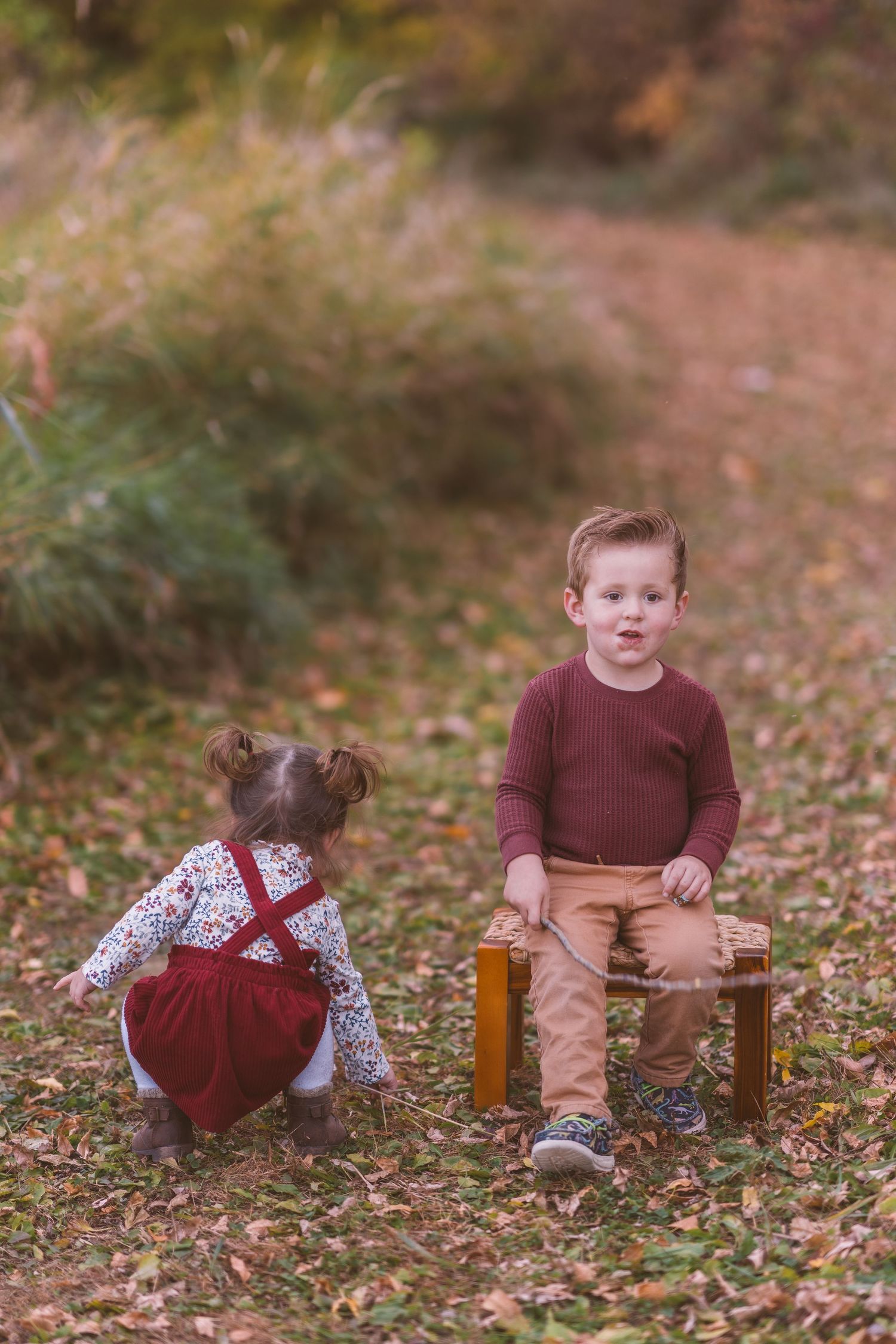 Two children in autumn outfits playing with wooden stool on fall path.