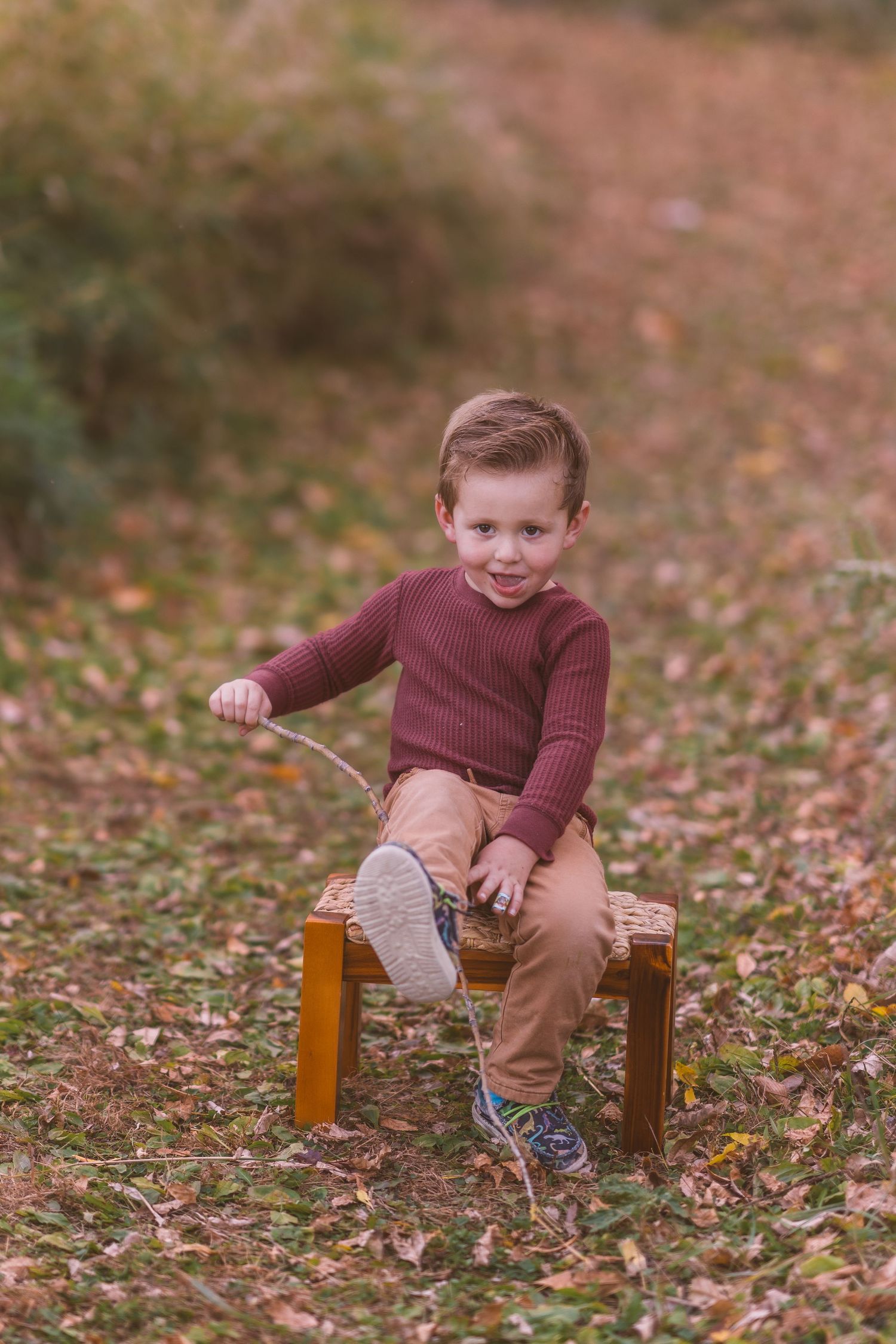 Child in brown sweater sits playfully on wooden stool surrounded by autumn leaves.